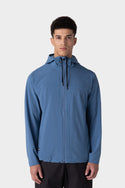 A young man stands facing forward against a plain light gray background, wearing the 686 Mens ATP Stretch Wind Jacket by 686 in light blue with a front zipper and hood.