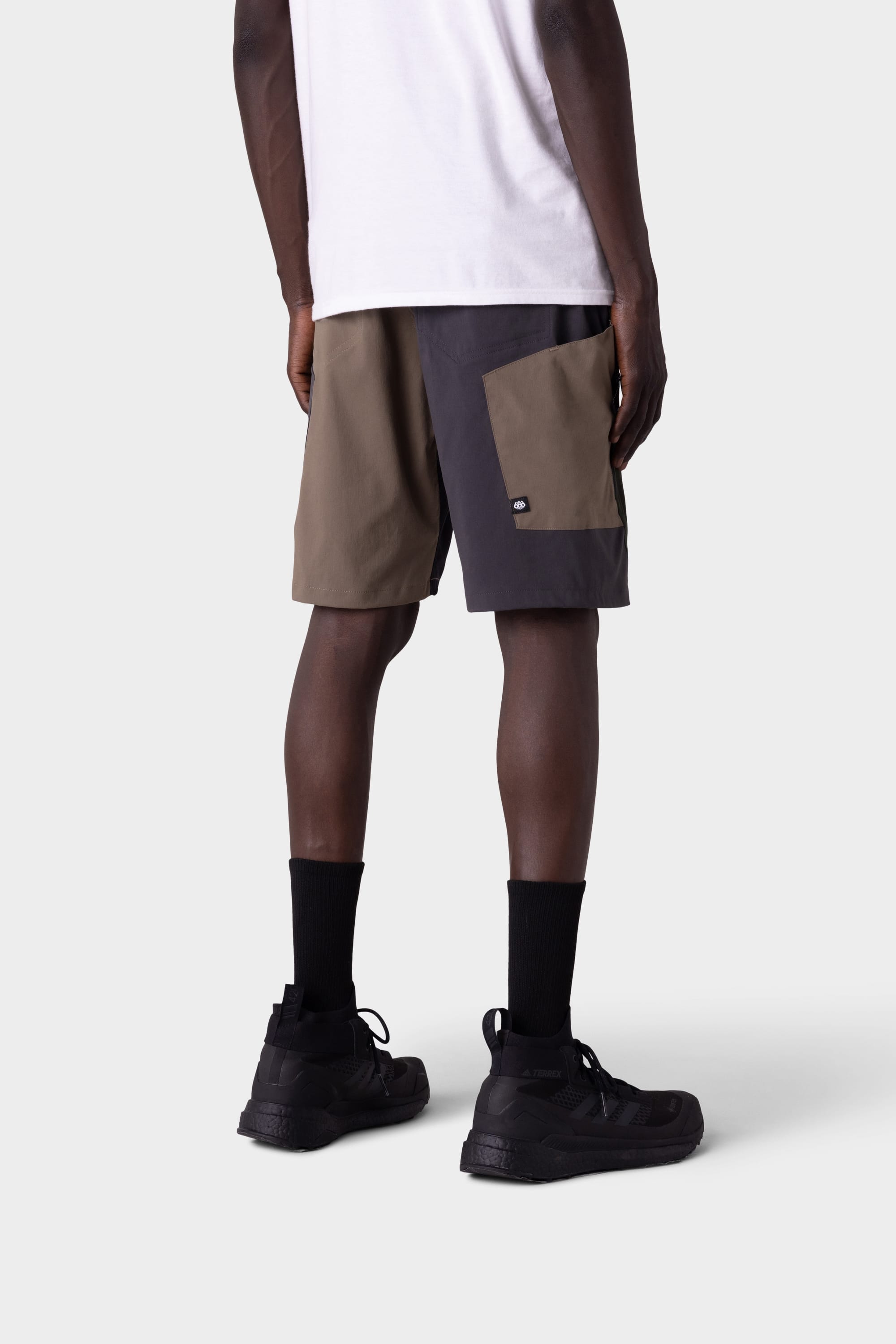 A person stands facing away, wearing a white t-shirt, 686 Mens Anything Hybrid Cargo Short by 686 in brown and dark gray, black crew socks, and black sneakers against a plain light gray background.