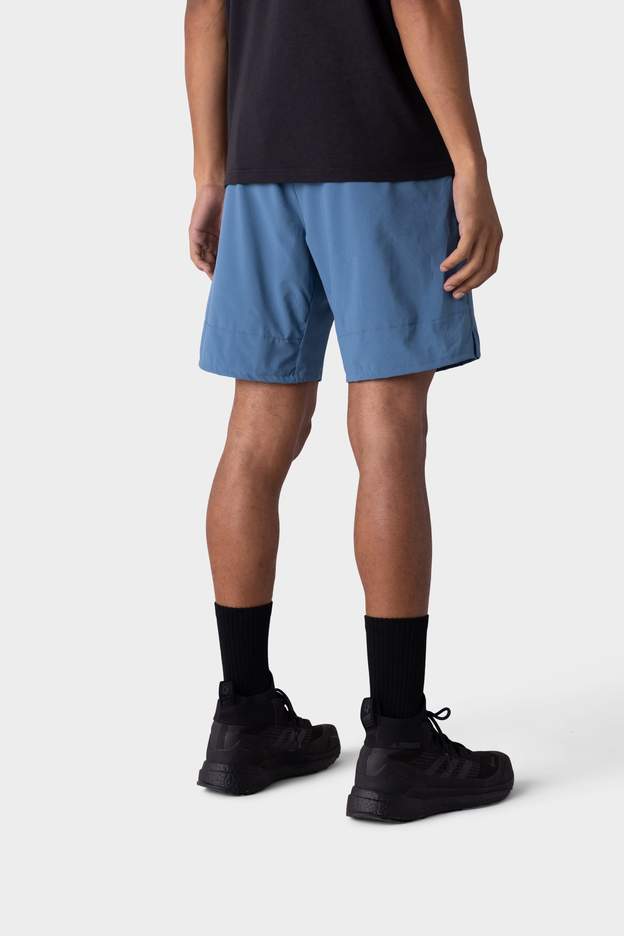 A person stands facing away in a black T-shirt, 686 Mens ATP Stretch Performance Shorts by 686, black crew socks, and black sneakers against a plain light background.