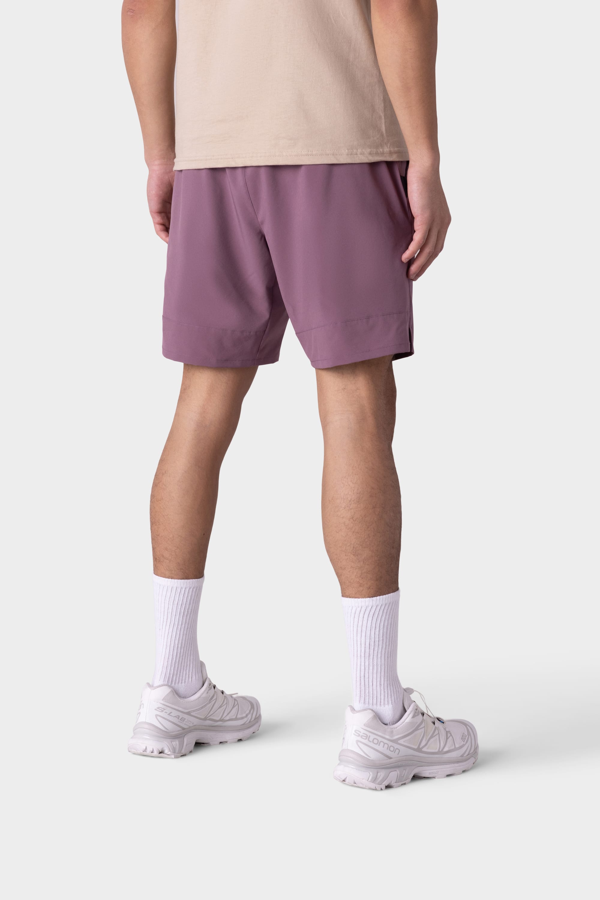 A person stands facing away, shown from mid-back down against a plain white background, wearing 686 Mens ATP Stretch Performance Short by 686 in mauve, paired with a beige T-shirt, white crew socks, and white sneakers.