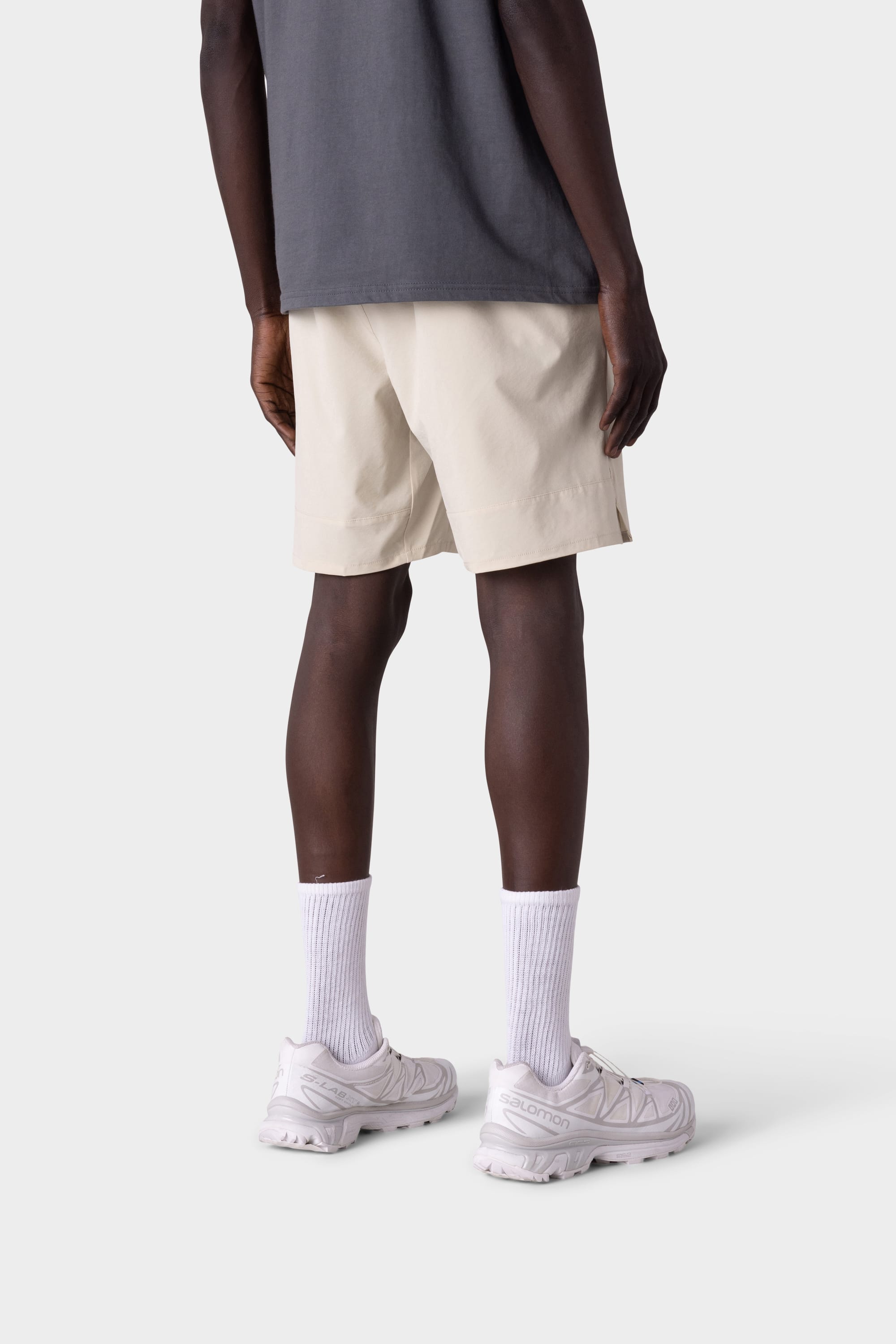 A person stands facing away in a dark gray shirt, 686 Mens ATP Stretch Performance Short by 686 in beige, white crew socks, and white athletic sneakers against a light gray background.