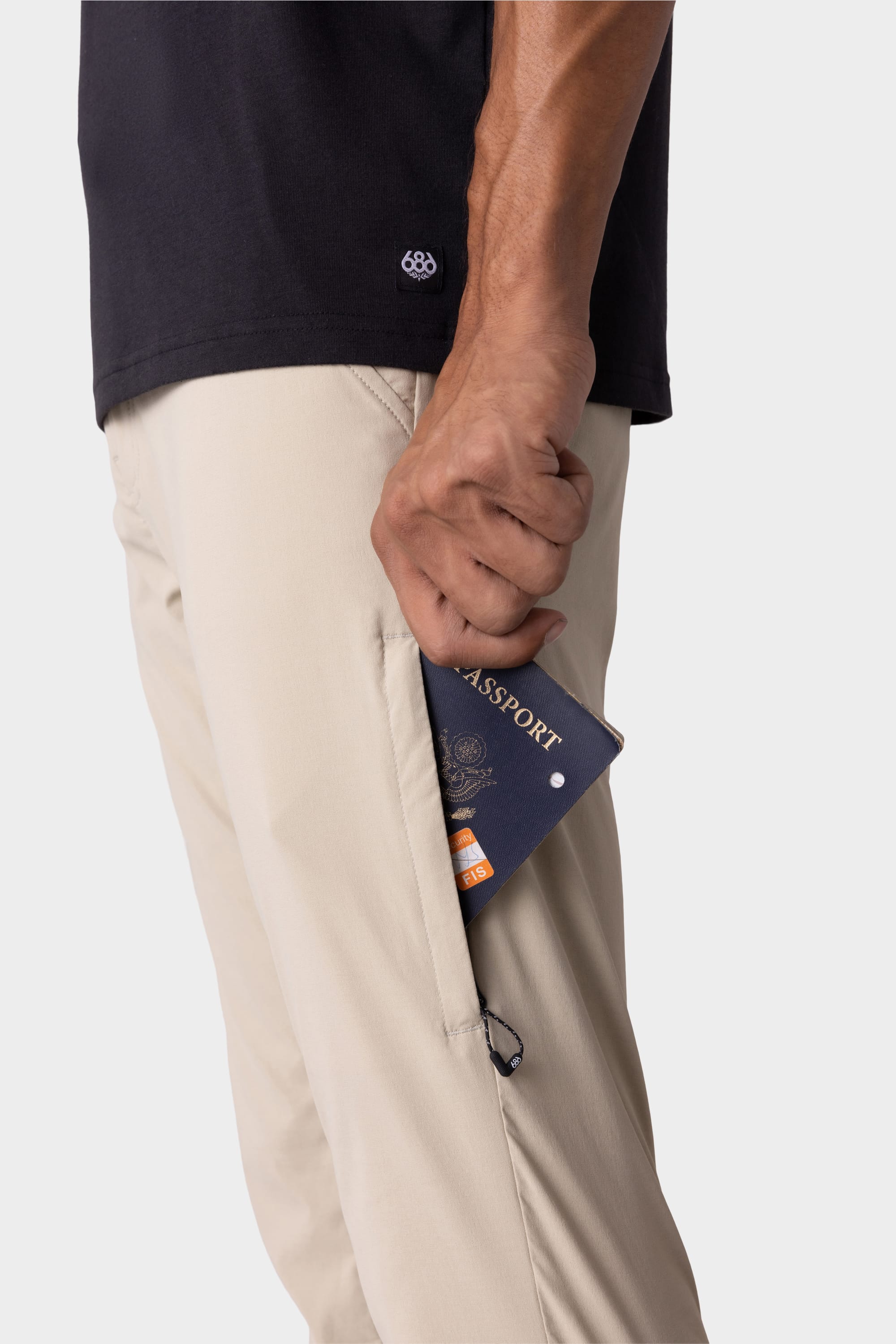 A person in 686 Mens Everywhere Featherlight Chino Pant - Slim Fit (beige) and a black shirt holds a navy blue passport partly tucked into the pants zippered side pocket—ideal for summer or warm weather adventures.