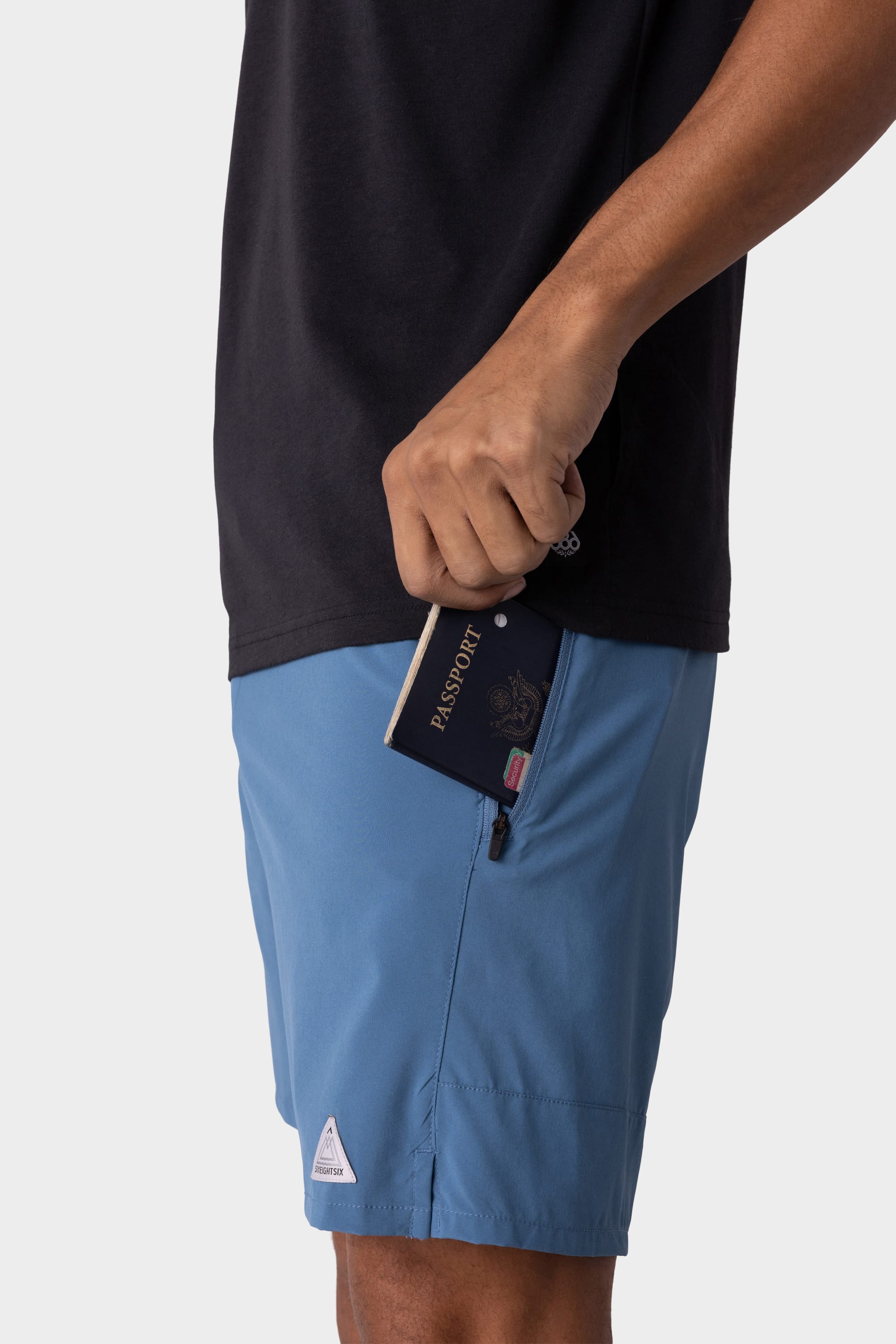 A person wearing a black shirt and 686 Mens ATP Stretch Performance Shorts is placing a passport into the zippered side pocket of their shorts.