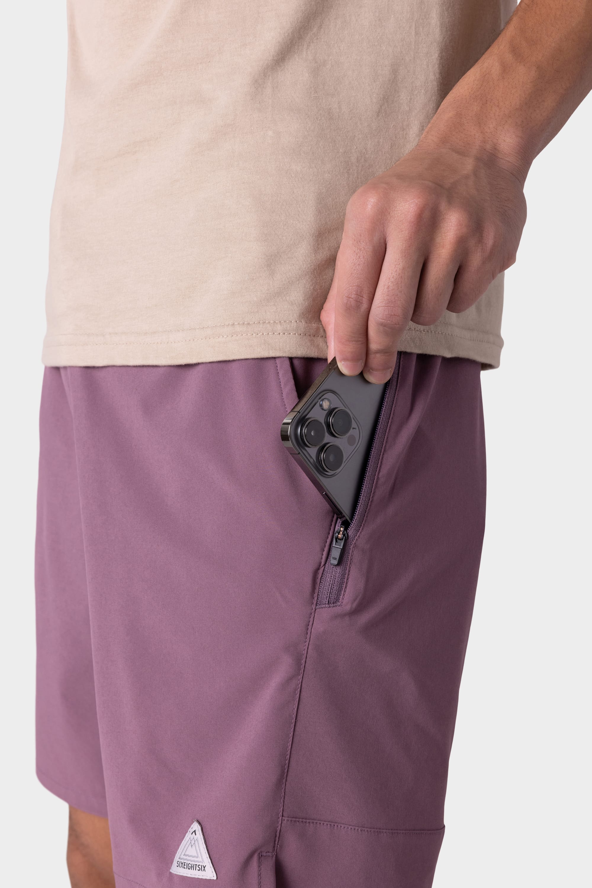 A person wearing a beige shirt and 686 Mens ATP Stretch Performance Short in mauve places a smartphone into the zippered side pocket of the shorts; only their torso and hand are visible.