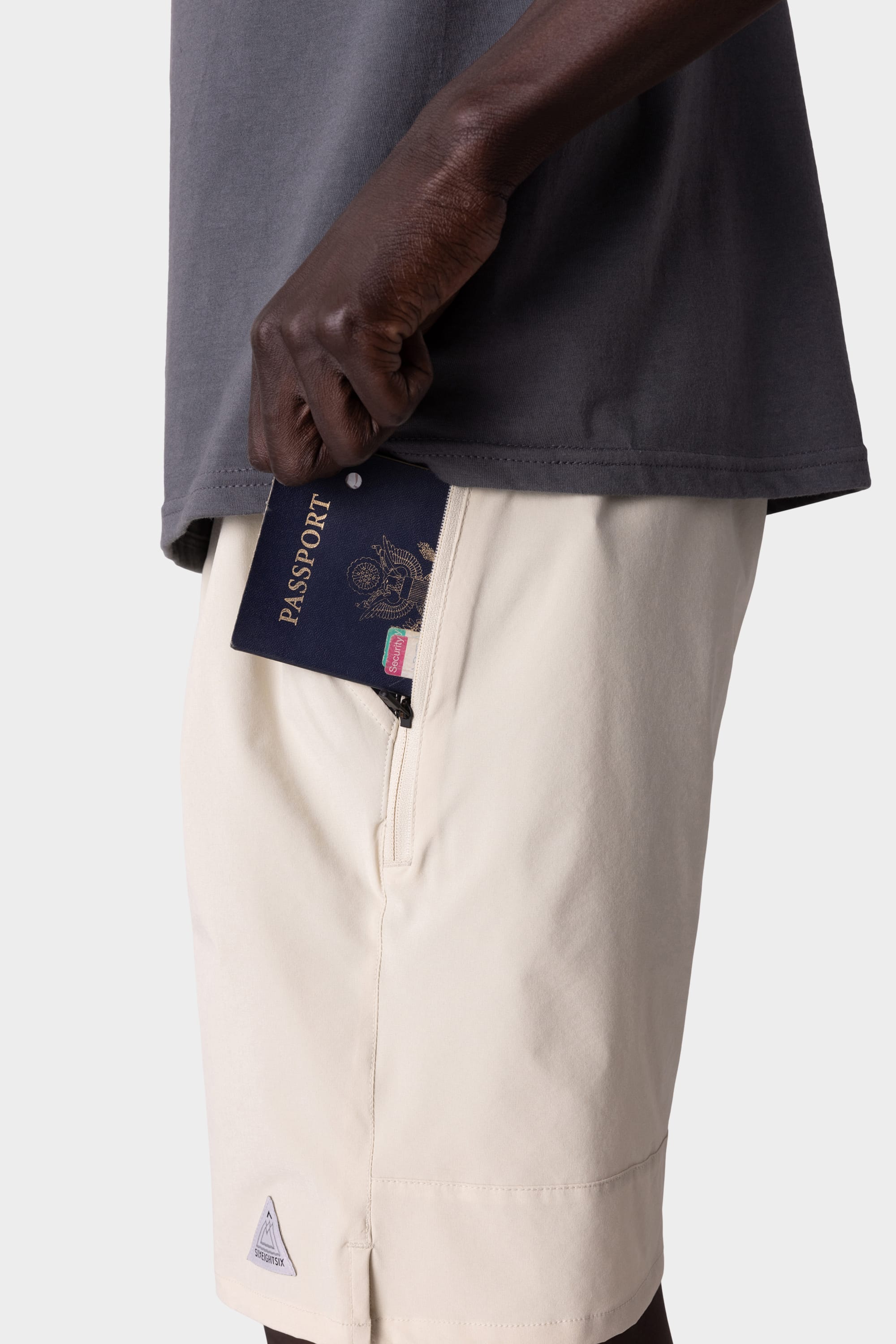 A person wearing 686 Mens ATP Stretch Performance Shorts in light color is placing a passport into the zippered pocket. The image, showing only their torso and hand, highlights the secure storage feature of the 686 shorts.