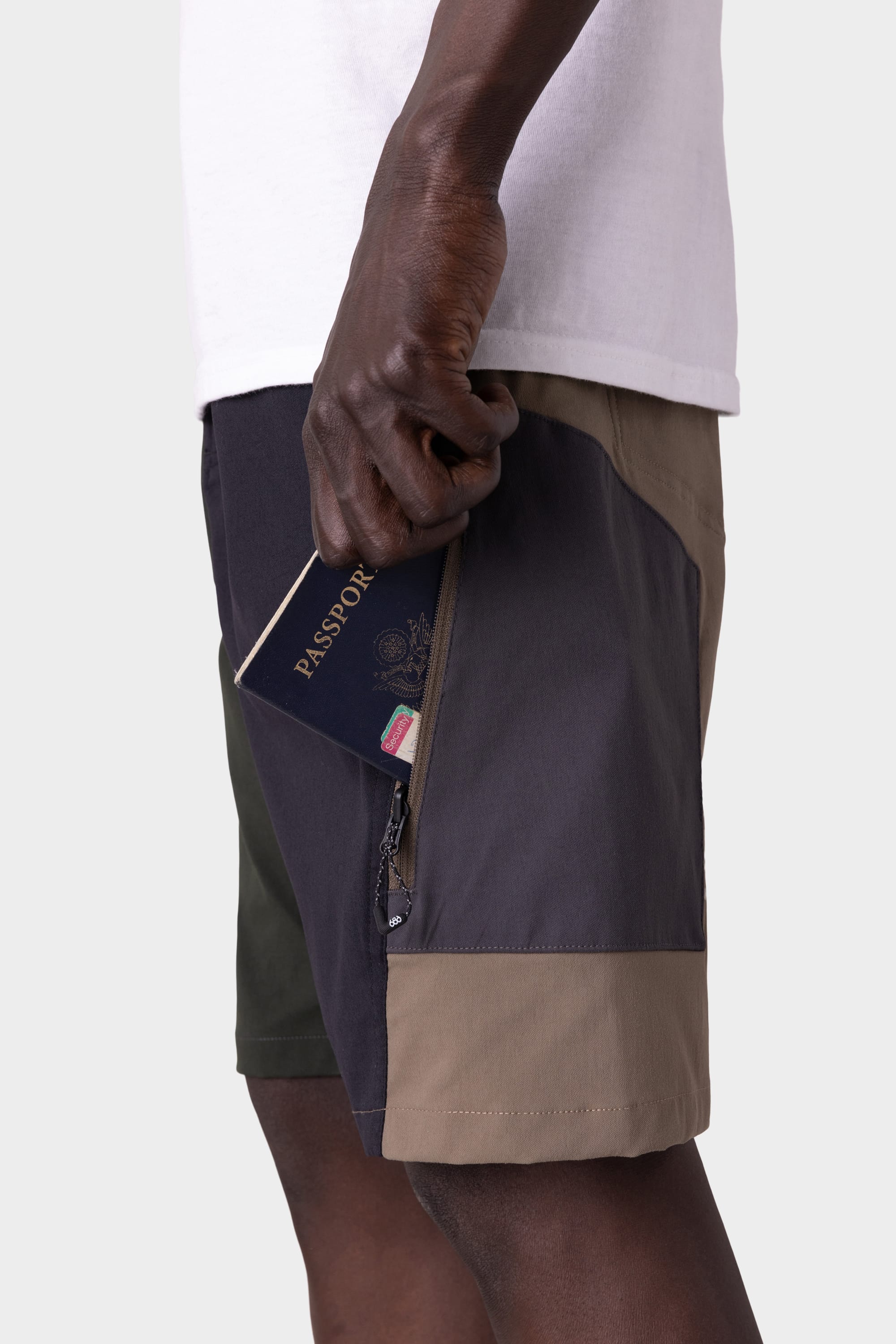 A man in 686 Mens Anything Hybrid Cargo Shorts by 686 slips a passport into the zippered side pocket. The minimalist waist-down shot features a white shirt and simple light backdrop, highlighting the color-block cargo style.
