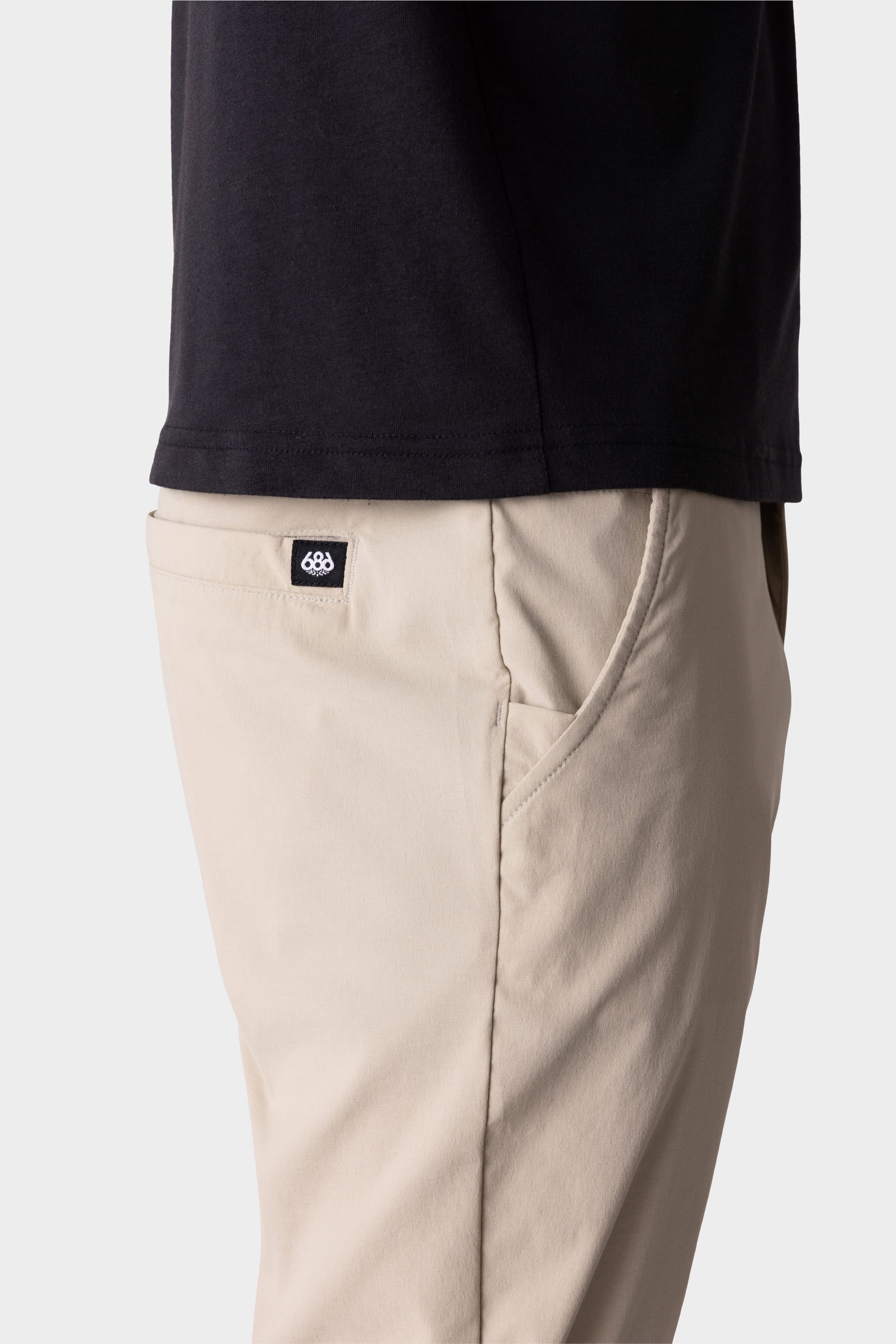 A person wears the 686 Mens Everywhere Featherlight Chino Pant - Slim Fit in beige, featuring a small black and white 686 logo tag above the pocket, paired with a black shirt. The image shows just the waist and upper thigh, ideal for warm weather.
