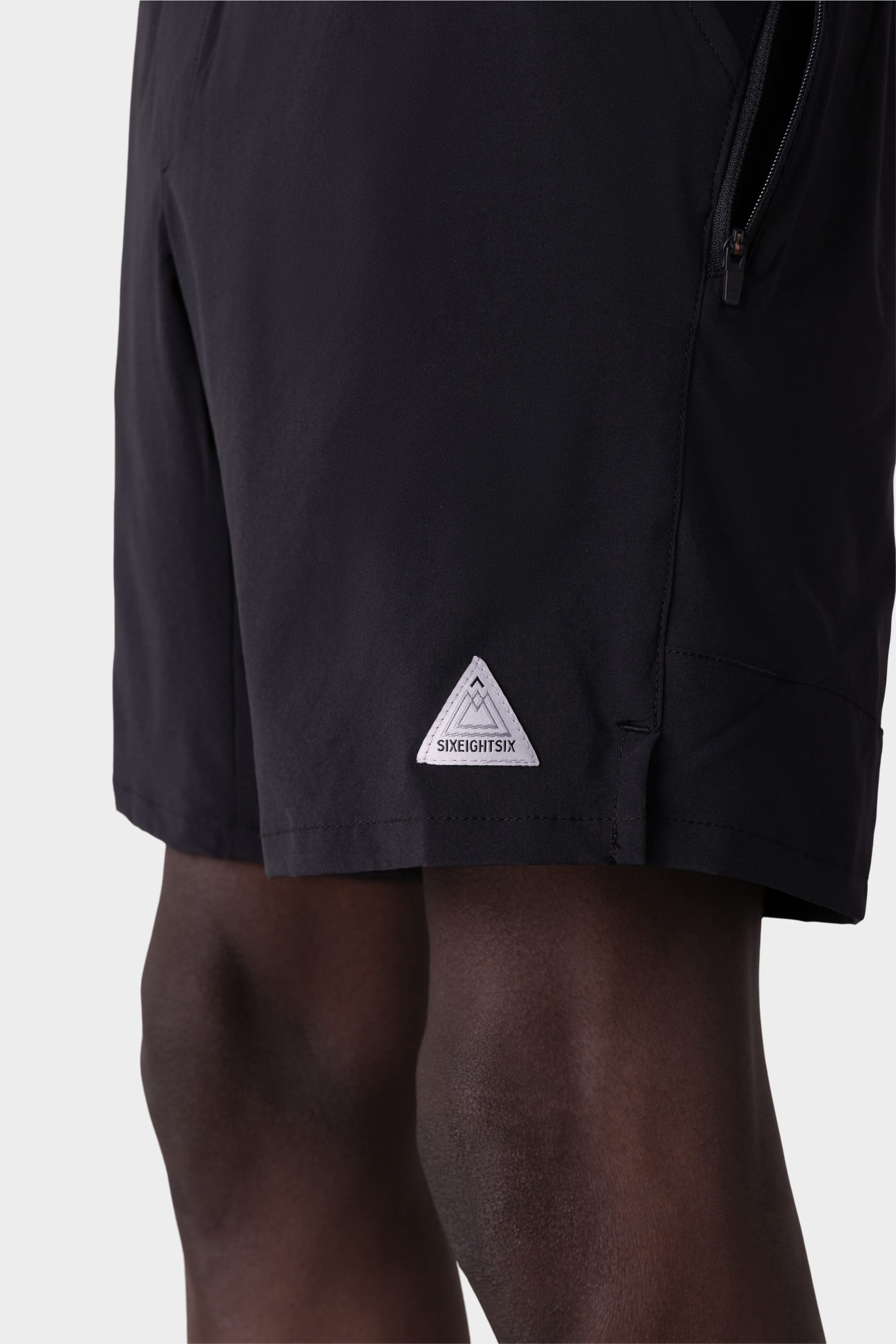 A close-up of a person wearing 686 Mens ATP Stretch Performance Shorts shows the black fabric, small white triangular 686 logo patch near the hem, part of their upper legs, and the shorts zipper pocket.