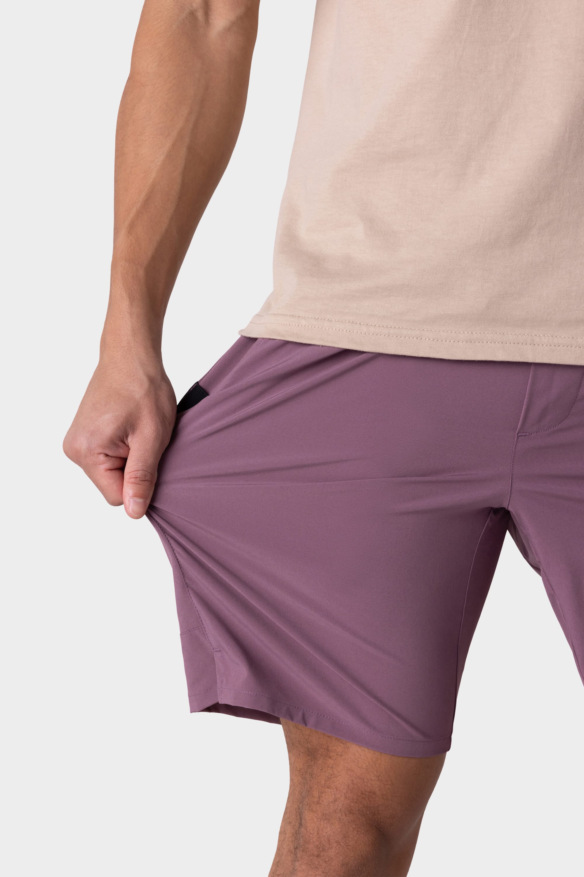 A person wearing a beige shirt and 686 Mens ATP Stretch Performance Shorts in mauve stretches the shorts outward to show their flexibility. Only the lower torso and upper legs are shown against a plain background.