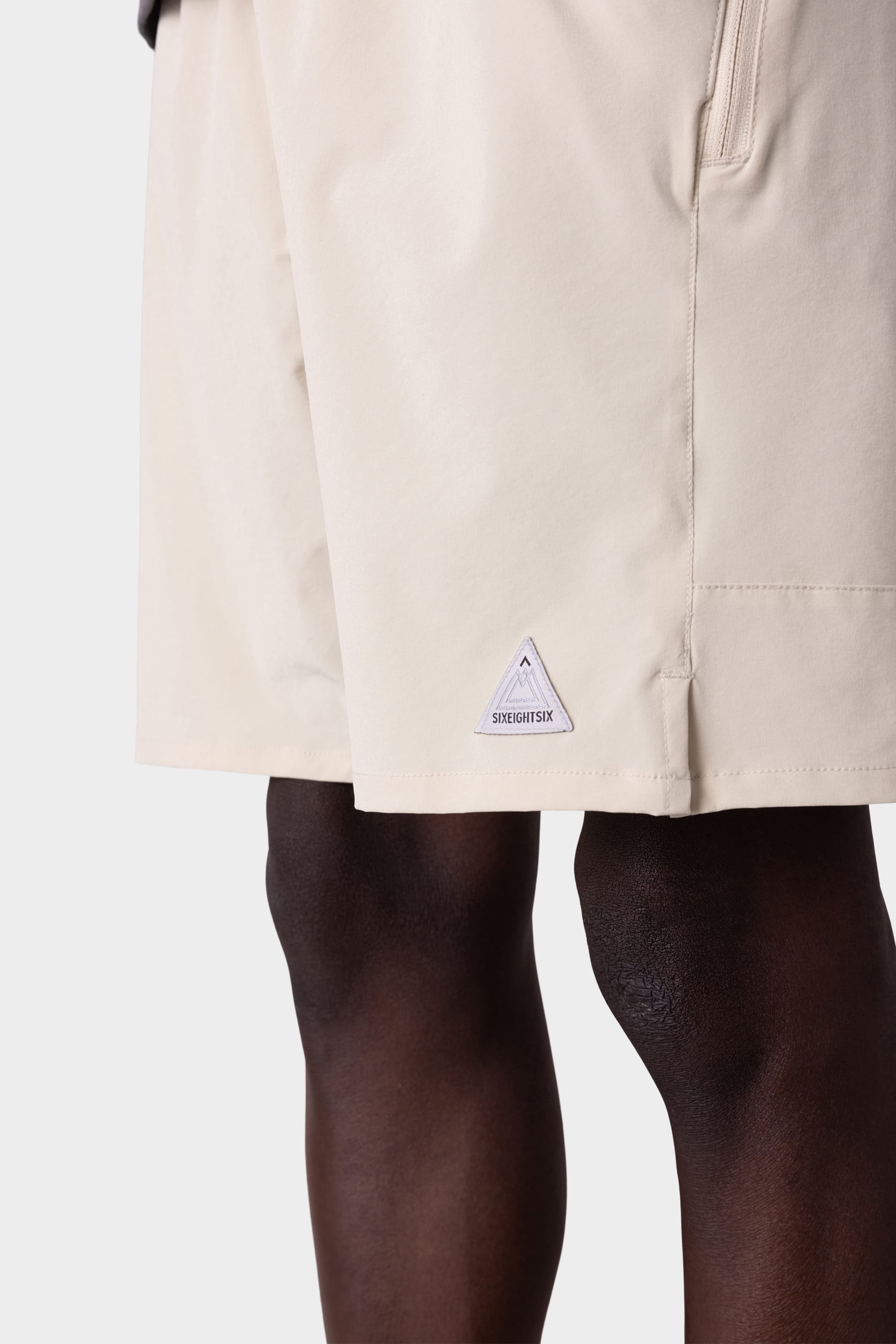 A close-up of a person wearing the 686 Mens ATP Stretch Performance Short in light beige, featuring a small triangular 686 logo near the hem, standing against a plain white background.