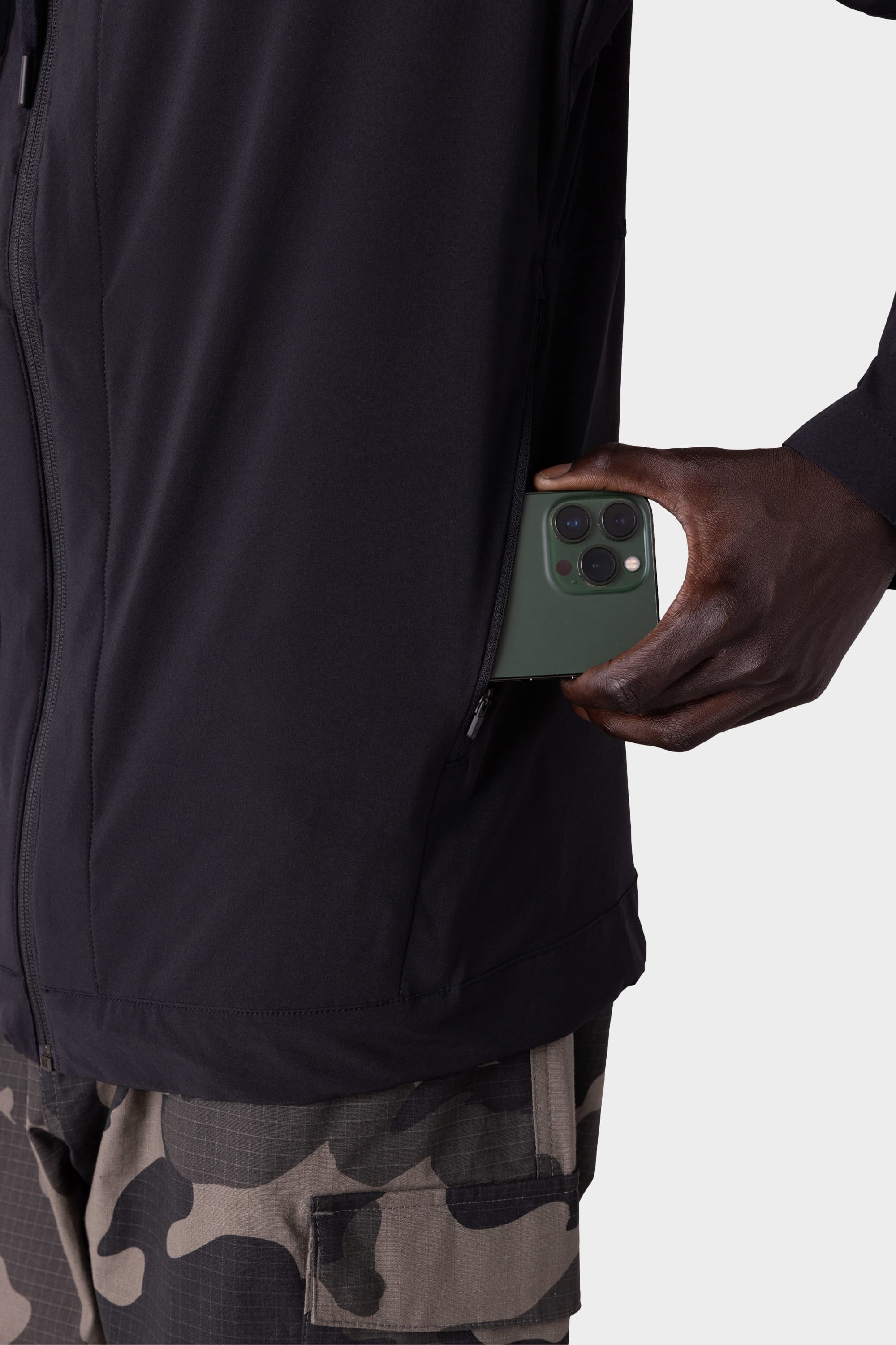 A person in the 686 Mens ATP Stretch Wind Jacket by 686 puts a green smartphone into its zippered pocket, set against a plain light background.