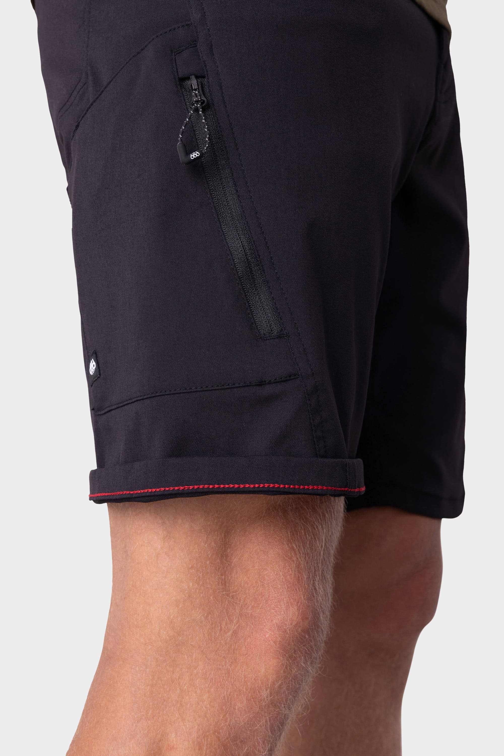 A close-up of the 686 Mens Anything Hybrid Cargo Short showcases its minimalist design, featuring dark fabric, a zippered pocket, and visible red stitching at the cuff. Only the shorts and lower thigh are visible.