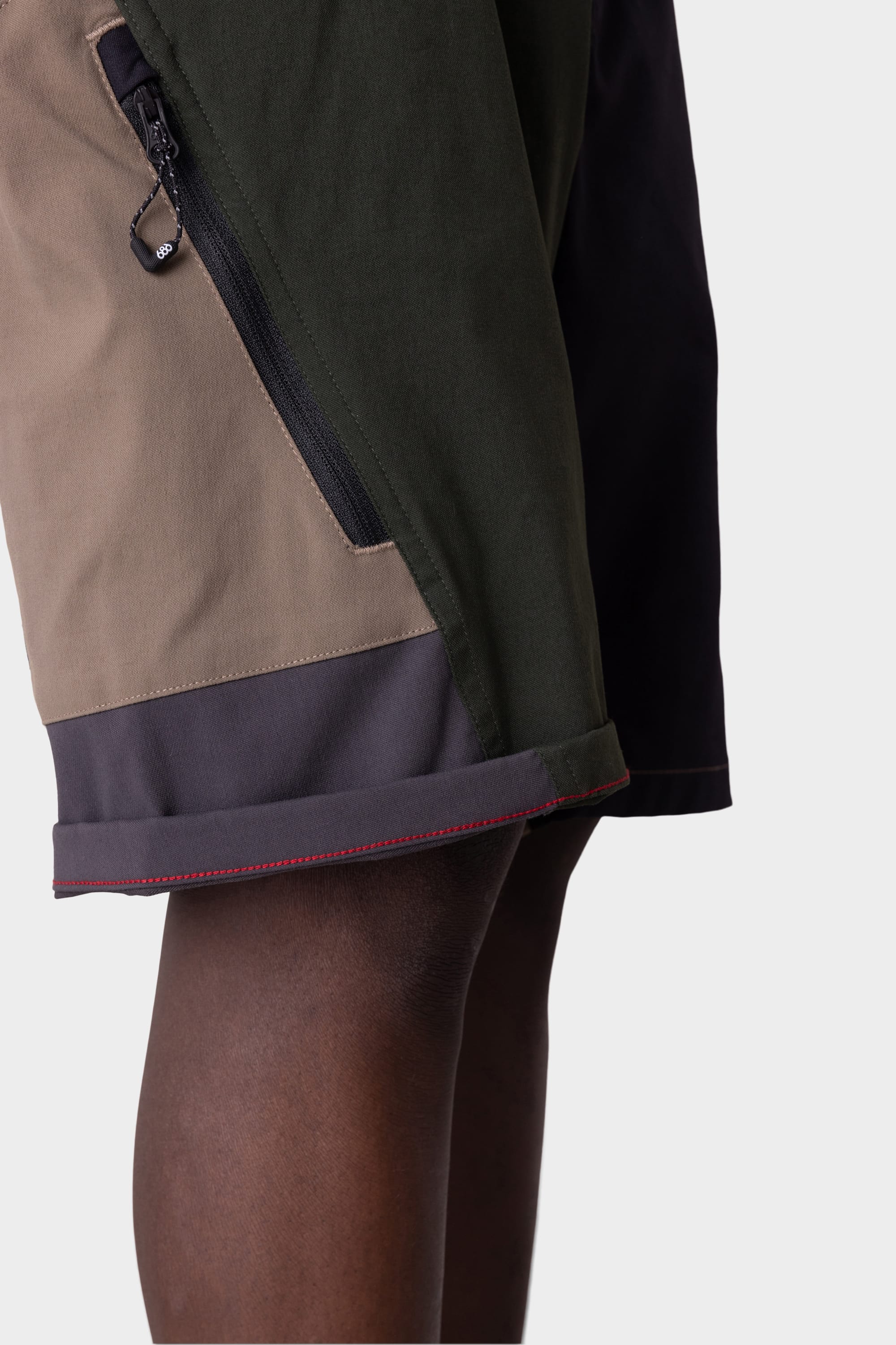A close-up of the 686 Mens Anything Hybrid Cargo Shorts by 686 in olive green, tan, and dark gray, featuring a minimalist design with a visible zipper pocket, shown against a plain light background.