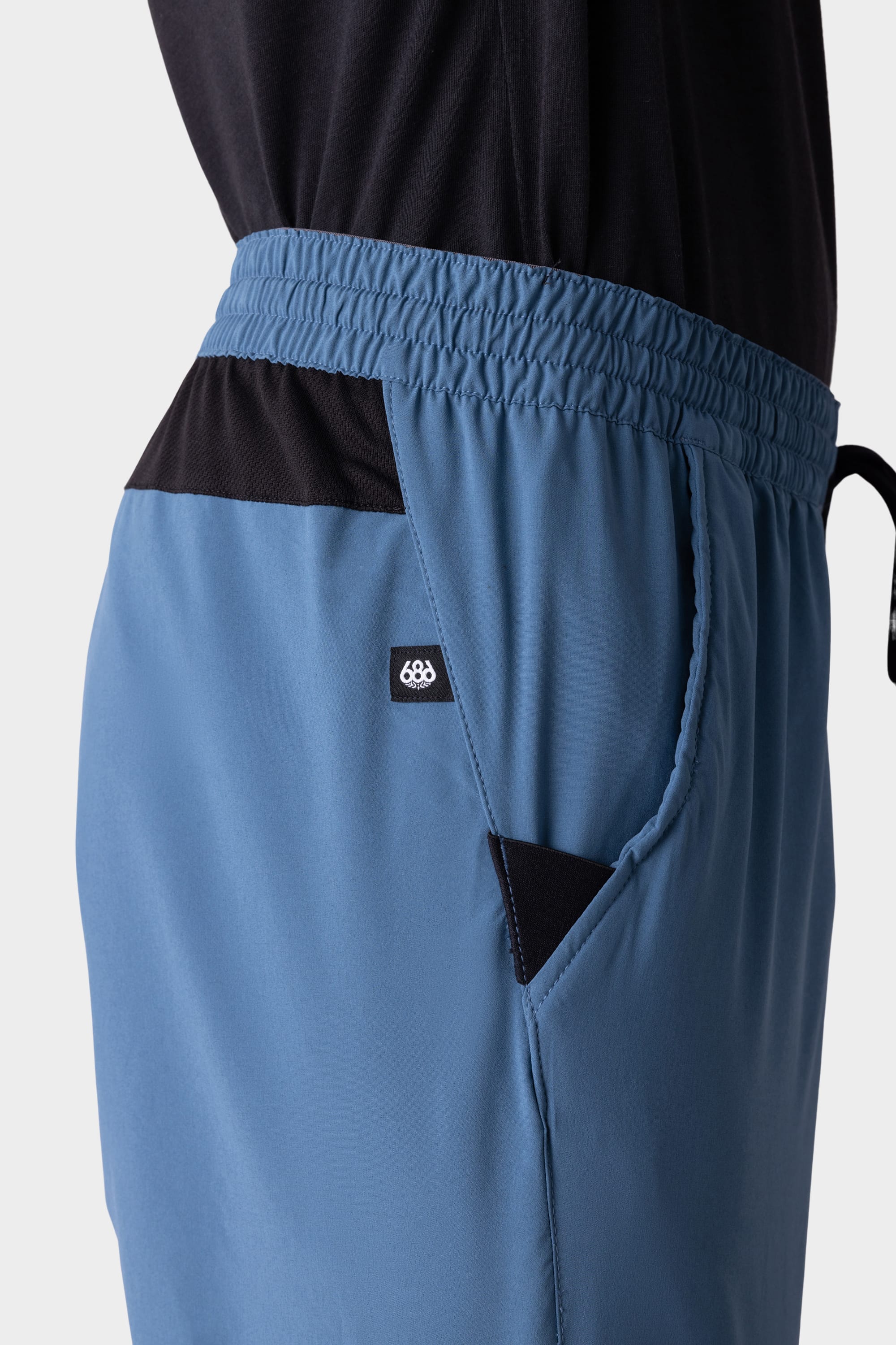 A close-up side view of a person wearing the 686 Mens ATP Stretch Performance Short, featuring blue fabric, a black waistband and pocket accents, plus a small 686 logo tag near the pocket.