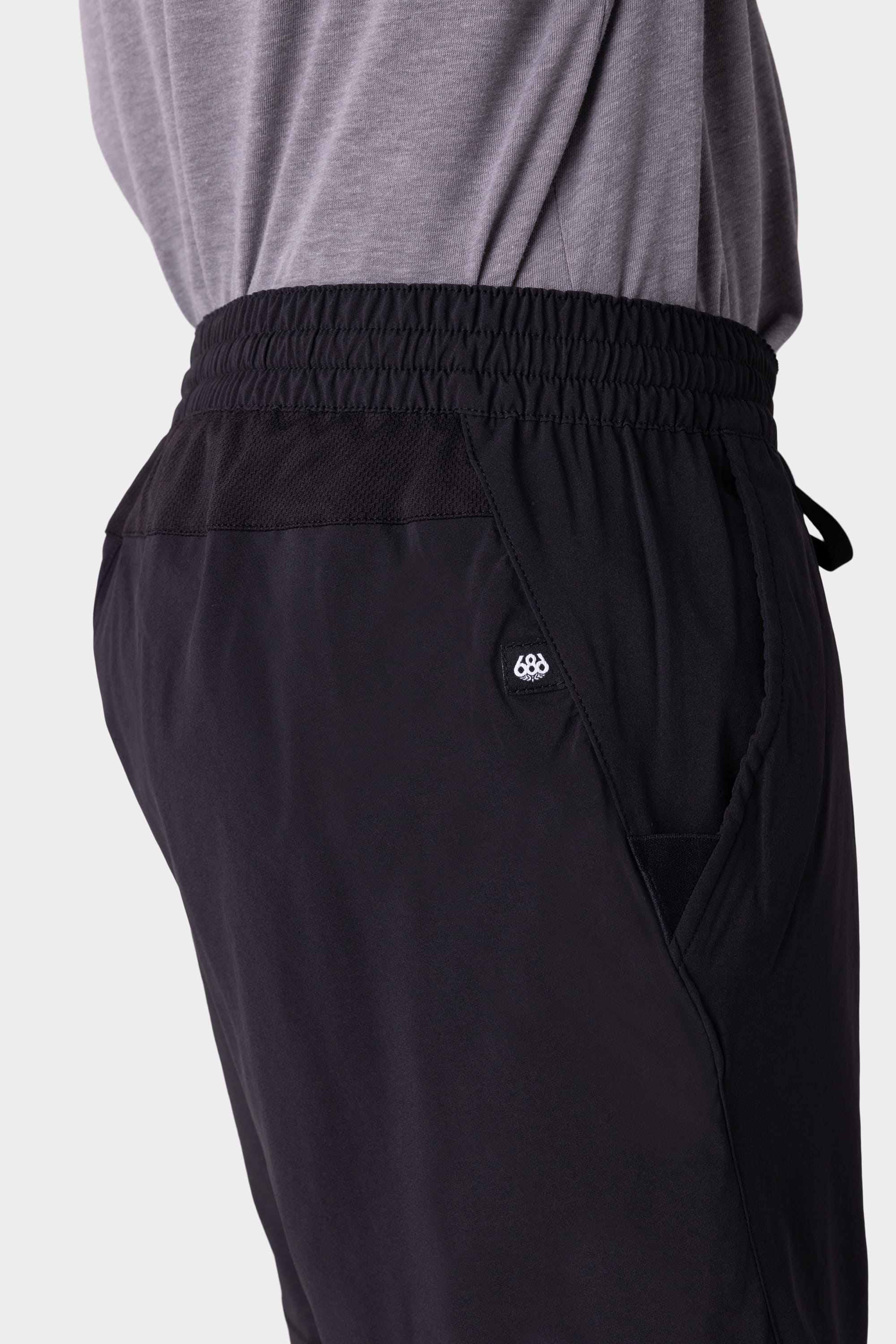 A close-up side view of a person wearing a grey shirt tucked into 686 Mens ATP Stretch Performance Shorts by 686, featuring black fabric, an elastic waistband, and a small circular logo near the pocket.