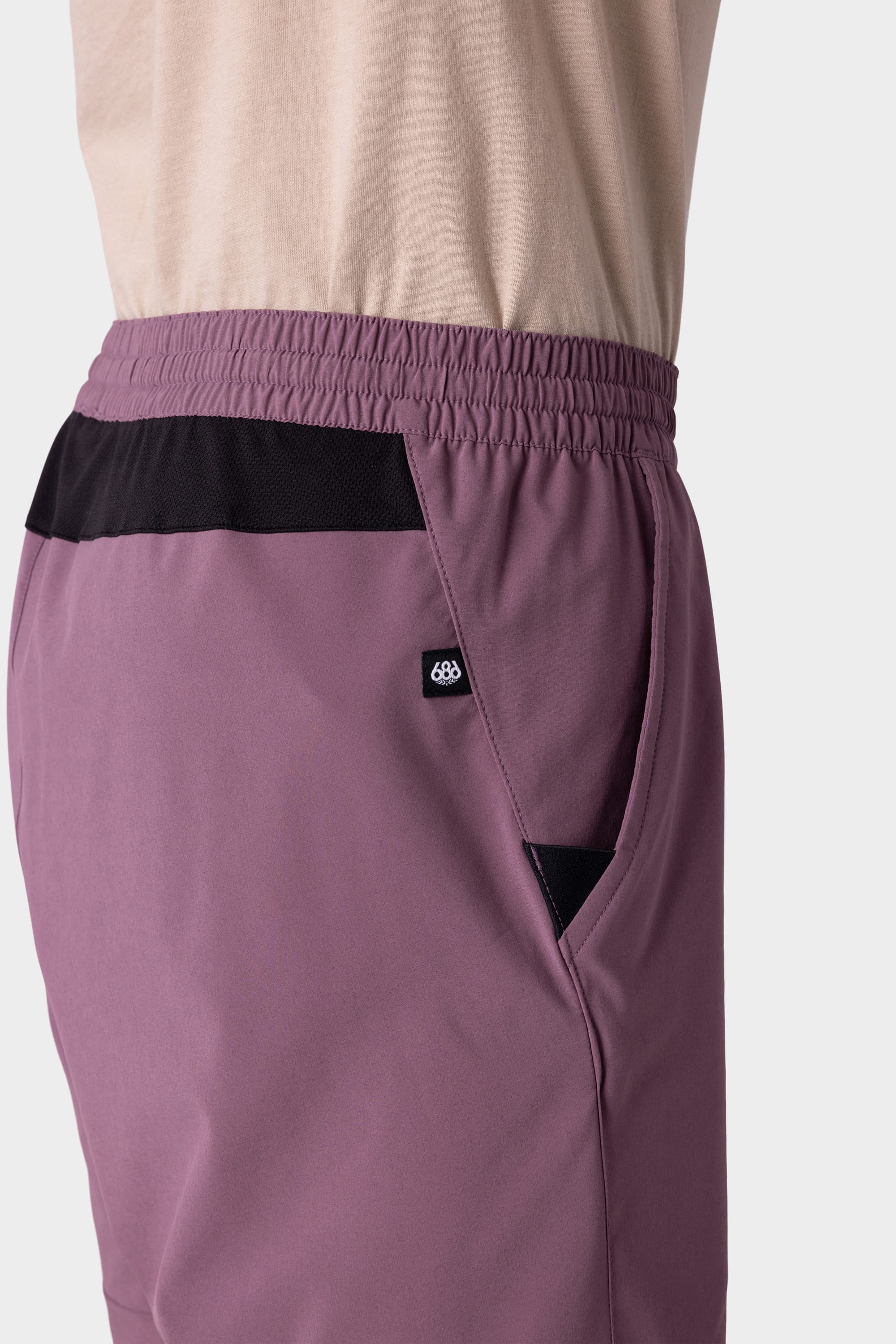 A person wears 686 Mens ATP Stretch Performance Shorts in mauve with a black pocket accent, elastic waistband, and round side logo, paired with a light beige shirt. Brand: 686.