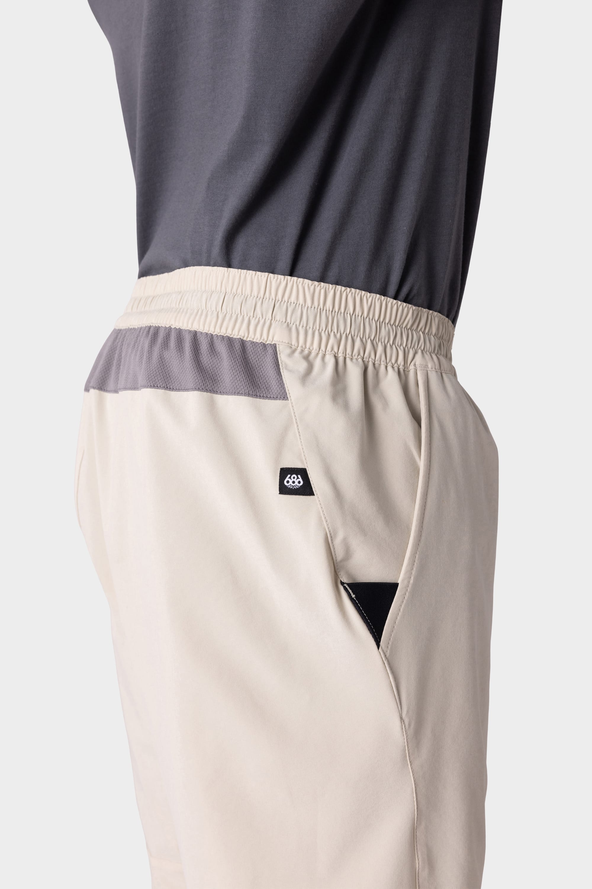 A person wears 686 Mens ATP Stretch Performance Shorts in light beige with a gray waistband and black and white logo detail near the pocket, paired with a dark gray shirt. Image is cropped to show only the lower torso and upper leg.