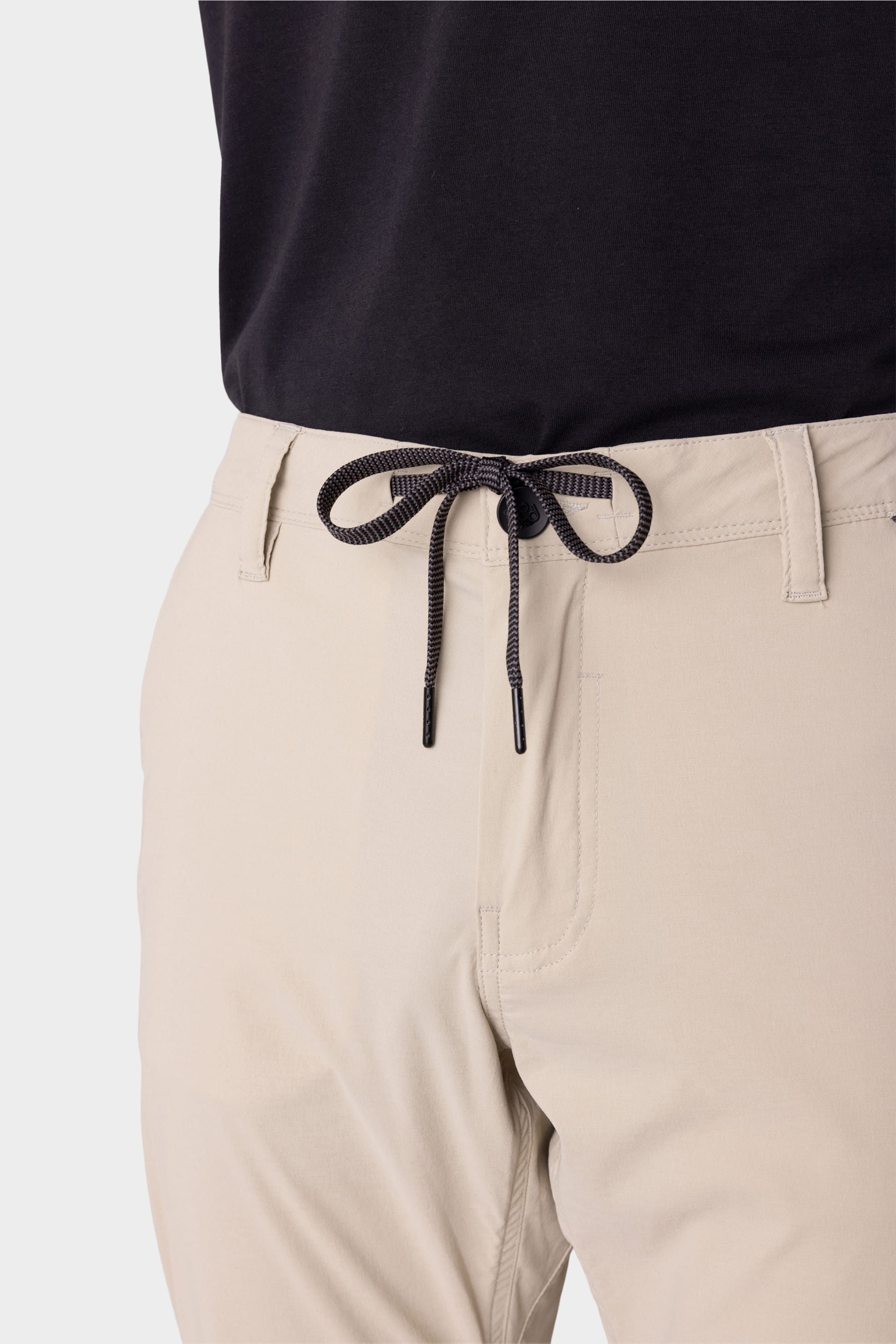 A close-up of a person wearing 686 Mens Everywhere Featherlight Chino Pant—slim fit, lightweight beige pants with a drawstring waist—paired with a tucked black shirt for stylish summer comfort from 686.