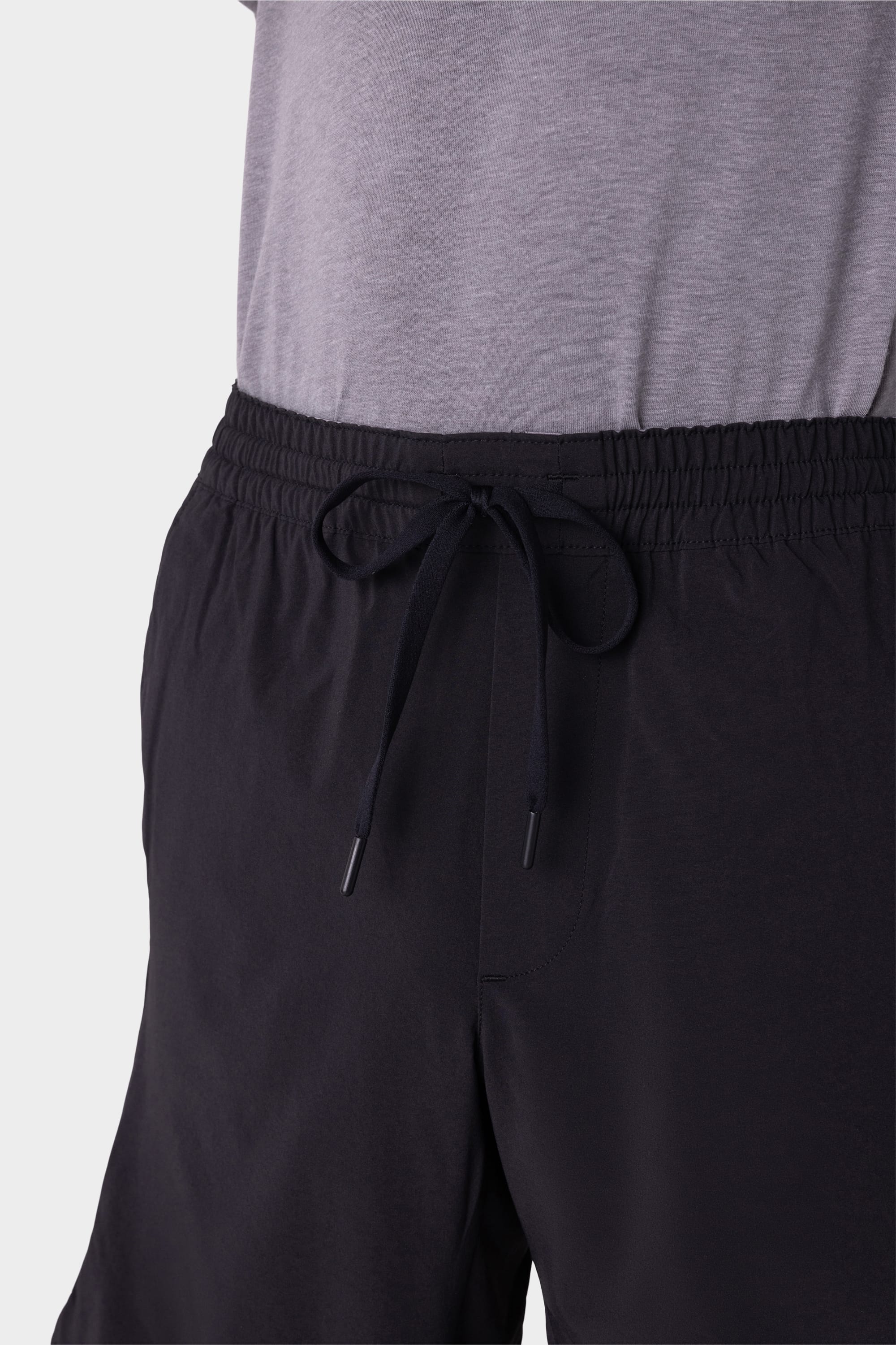 Close-up of a person wearing 686 Mens ATP Stretch Performance Shorts with a visible waistband and tied drawstring at the center, paired with a light grey shirt. The background is plain and light-colored.