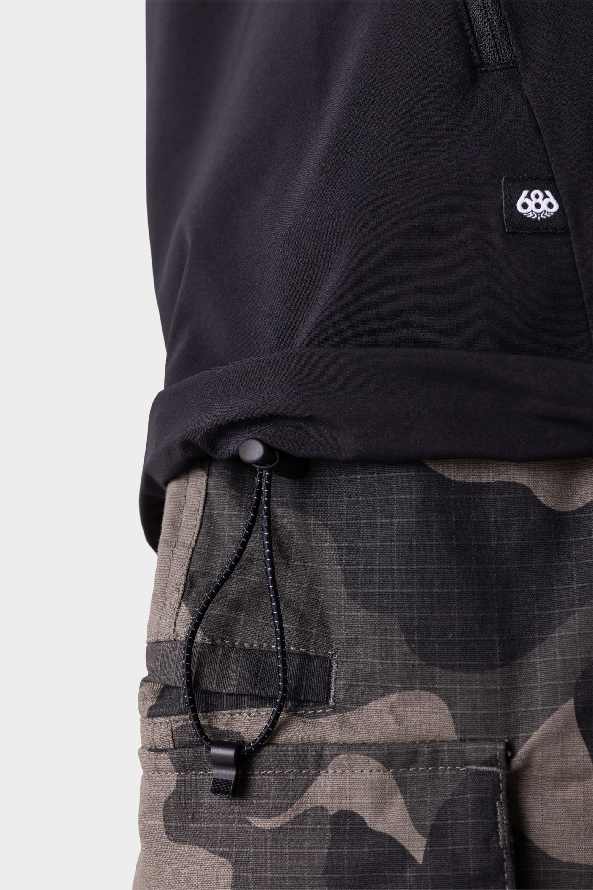 Close-up of a person wearing the 686 Mens ATP Stretch Wind Jacket with drawstring hem and gray camouflage cargo pants. The 686 logo is visible on the jacket. Only the upper body and waist are shown.