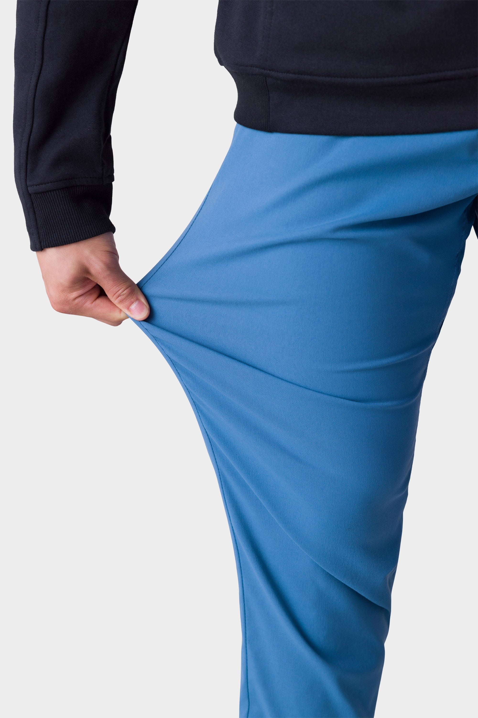 A person wearing 686 Mens Everywhere Merino-Lined Pant - Slim Fit stretches the flexible blue fabric with their left hand, pairing it with a black long-sleeve top against a plain, light background—ideal for colder conditions.