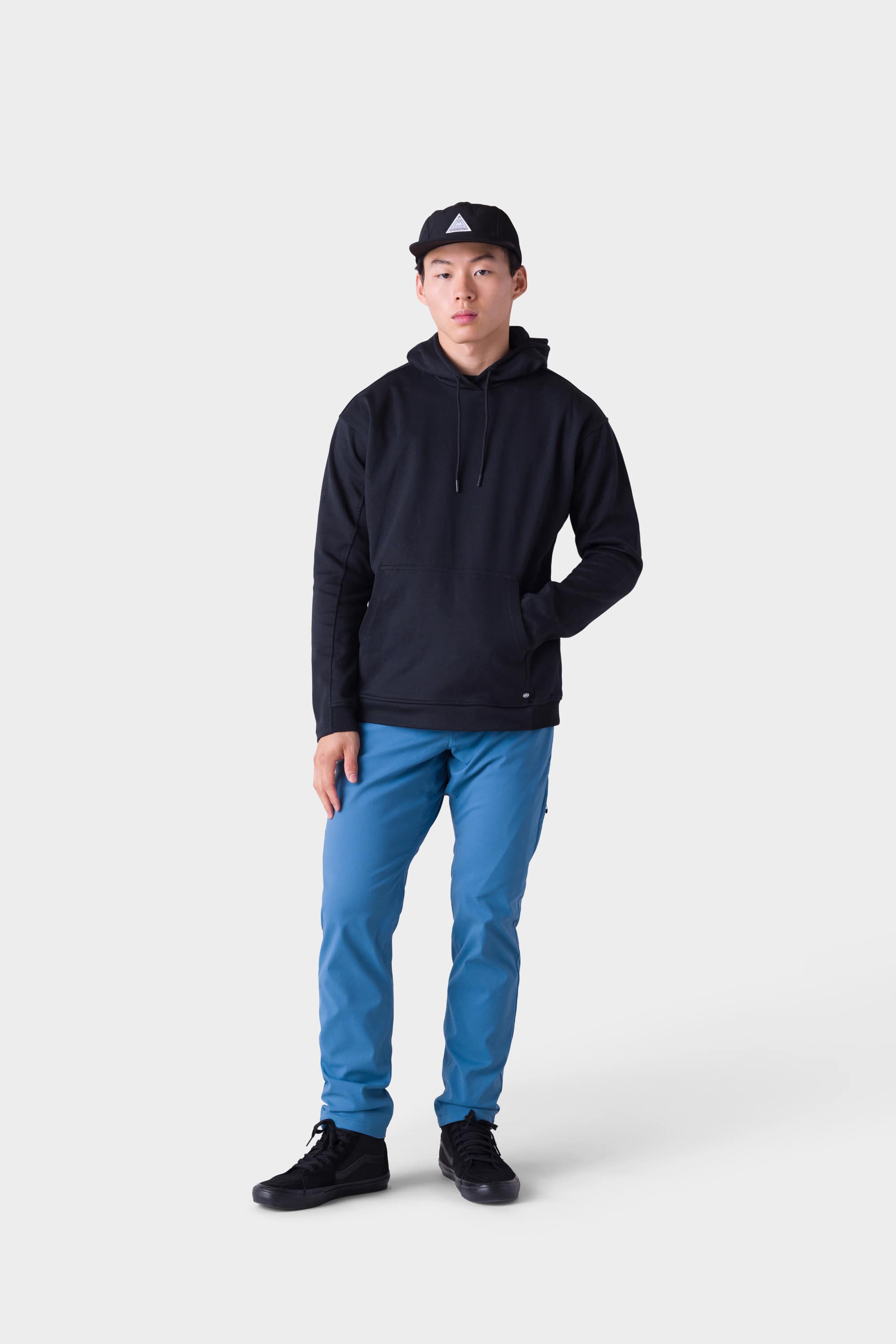 A person stands against a light gray background wearing the 686 Mens Everywhere Merino-Lined Pant - Slim Fit, paired with a black hoodie, black sneakers, and a black cap. One hand is in their pocket, and they have a neutral expression.