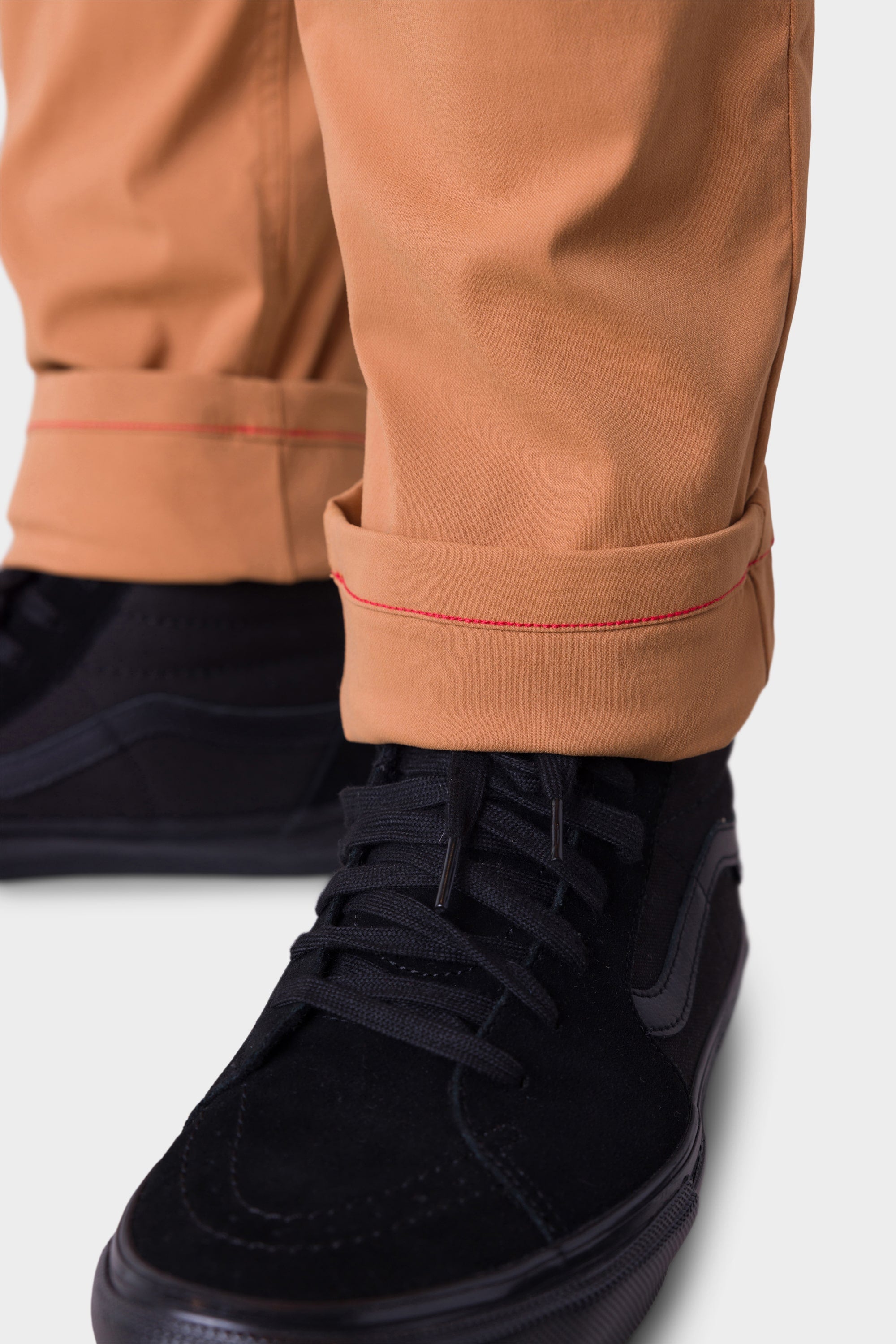 A close-up of the lower legs and feet shows someone wearing 686 Men’s Everywhere Merino-Lined Pant - Slim Fit in tan, with red-stitched rolled cuffs, paired with black lace-up sneakers—a stylish way to stay warm in colder conditions.