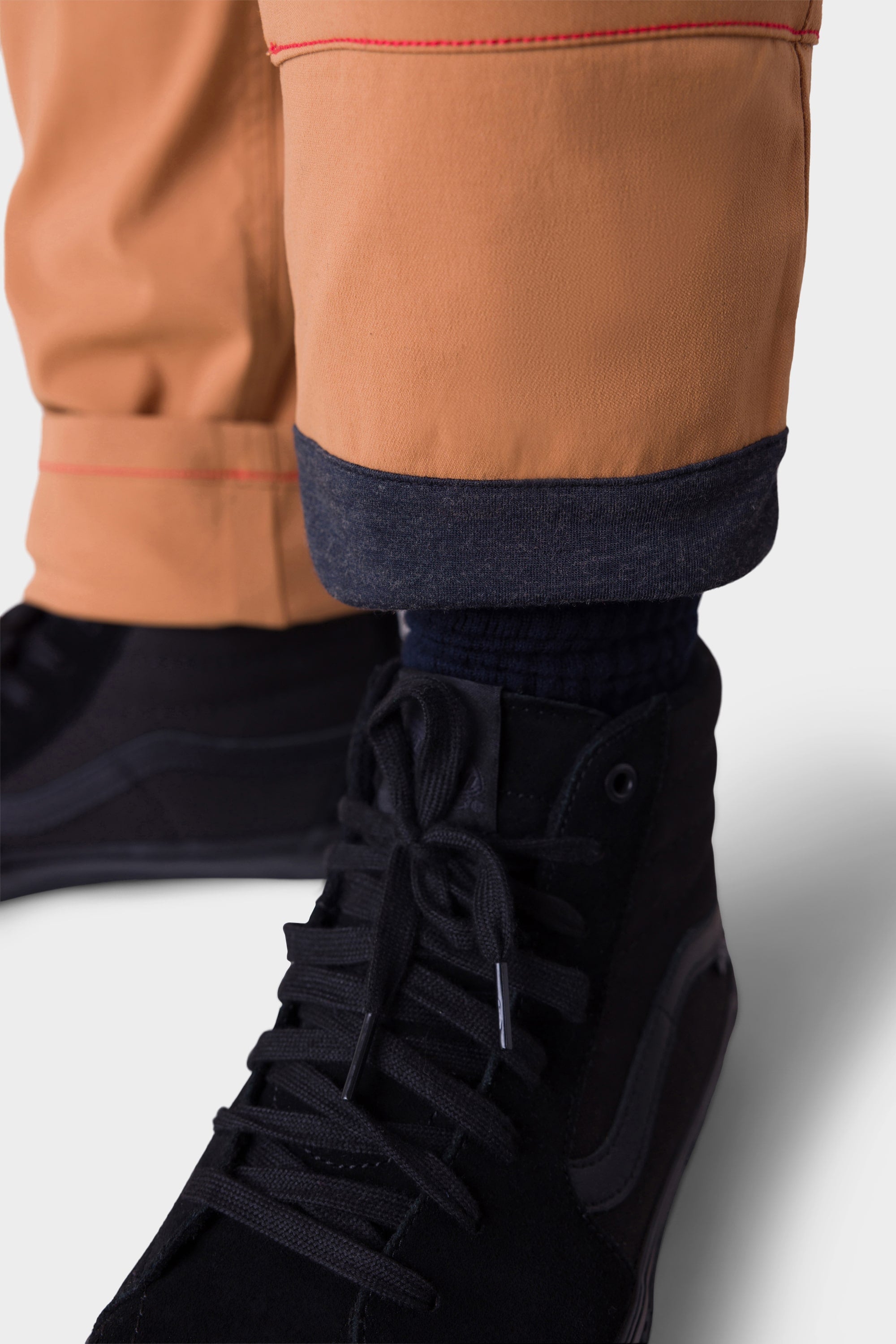 Close-up of a person wearing 686 Mens Everywhere Merino-Lined Pant - Slim Fit in tan, featuring rolled cuffs that reveal the dark denim lining. Styled with black sneakers, these pants by 686 are ideal for warmth in cold weather against a plain light background.