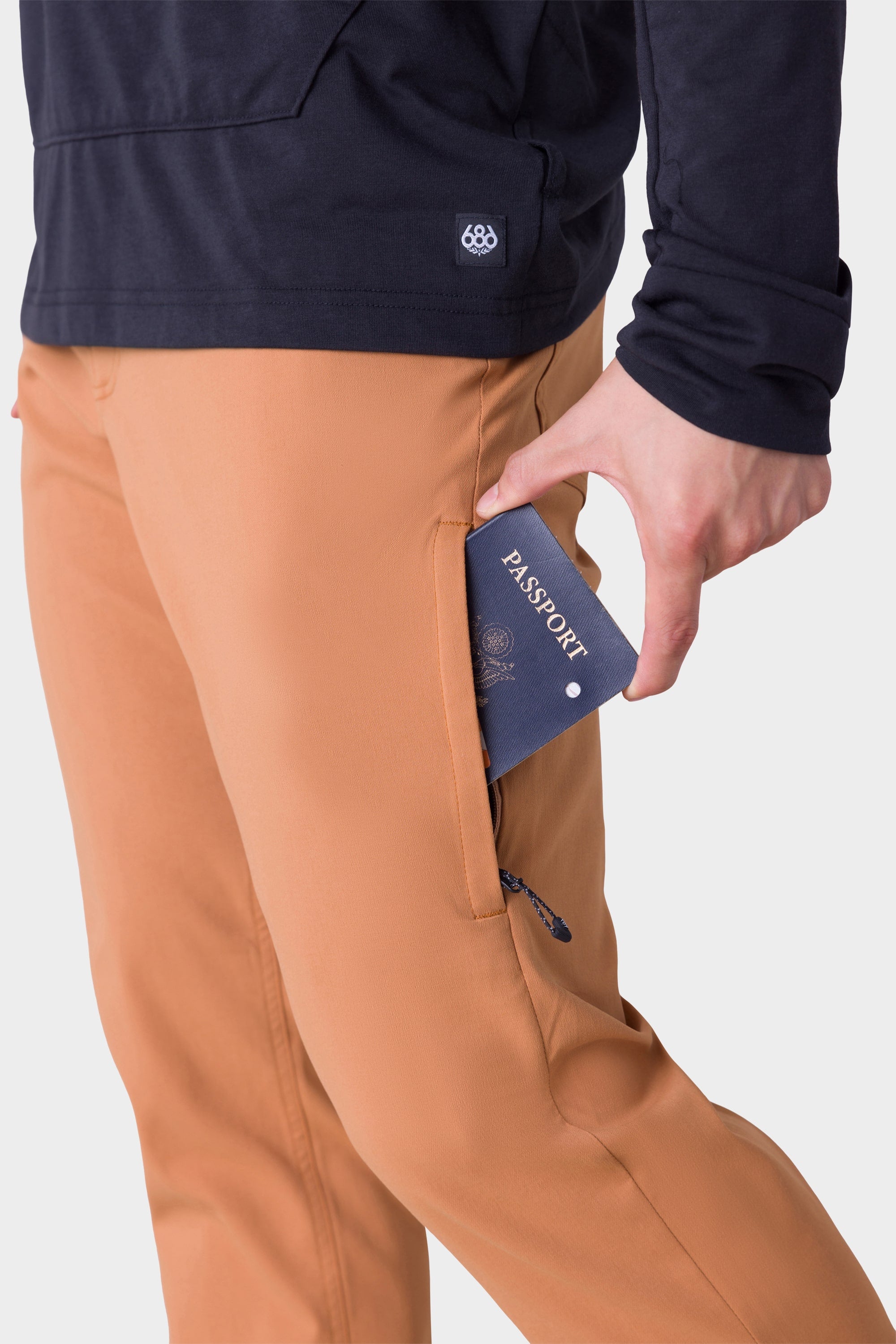 A person wearing 686 Mens Everywhere Merino-Lined Pant - Slim Fit in tan slips a blue passport into the zippered side pocket. The image highlights the secure pocket and warmth of these 686 pants designed for cold, wet conditions.