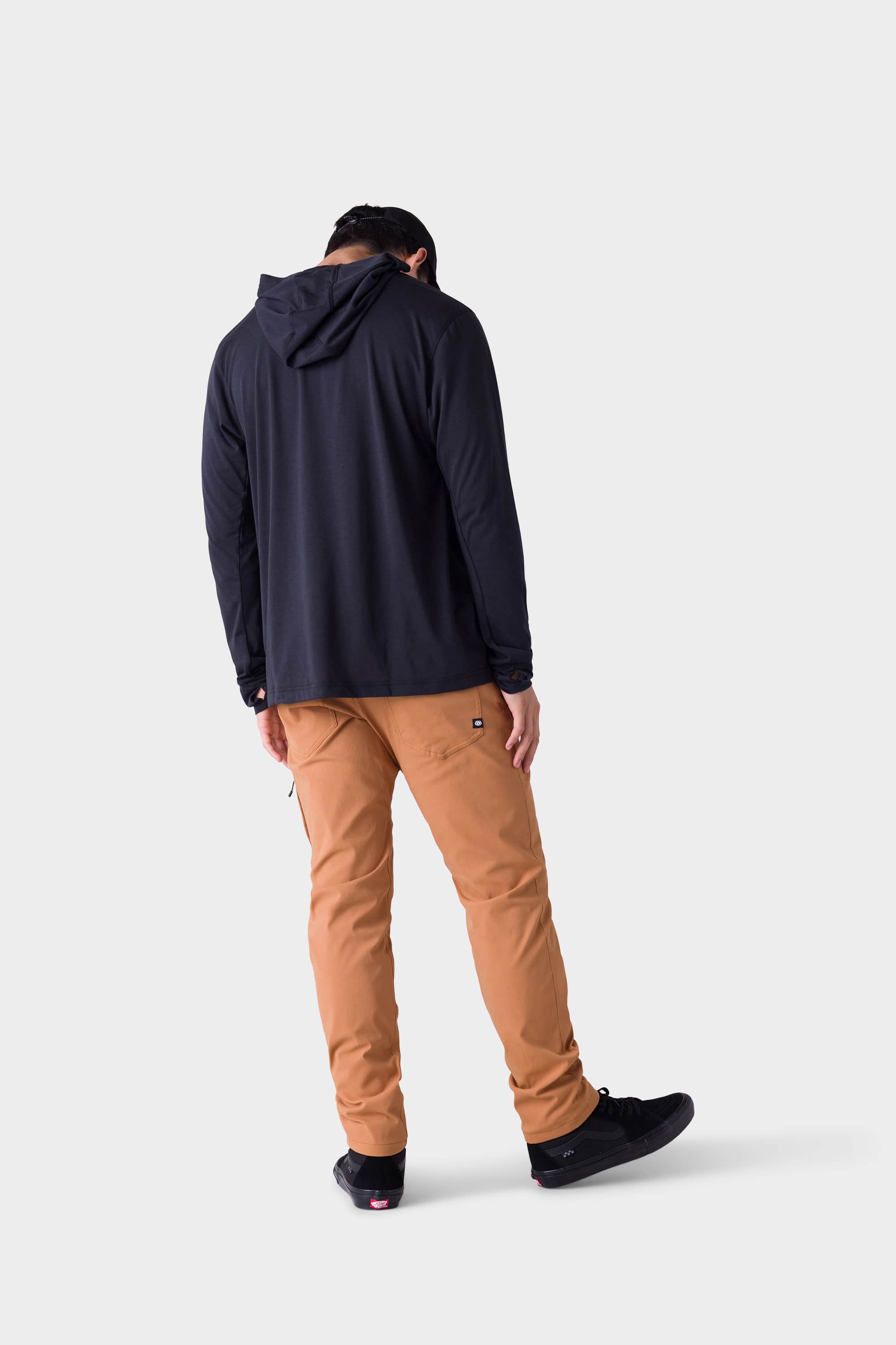 A person stands facing away against a plain light gray background, wearing black sneakers, a black hooded long-sleeve shirt, and 686 Mens Everywhere Merino-Lined Pant - Slim Fit by 686 in light brown.
