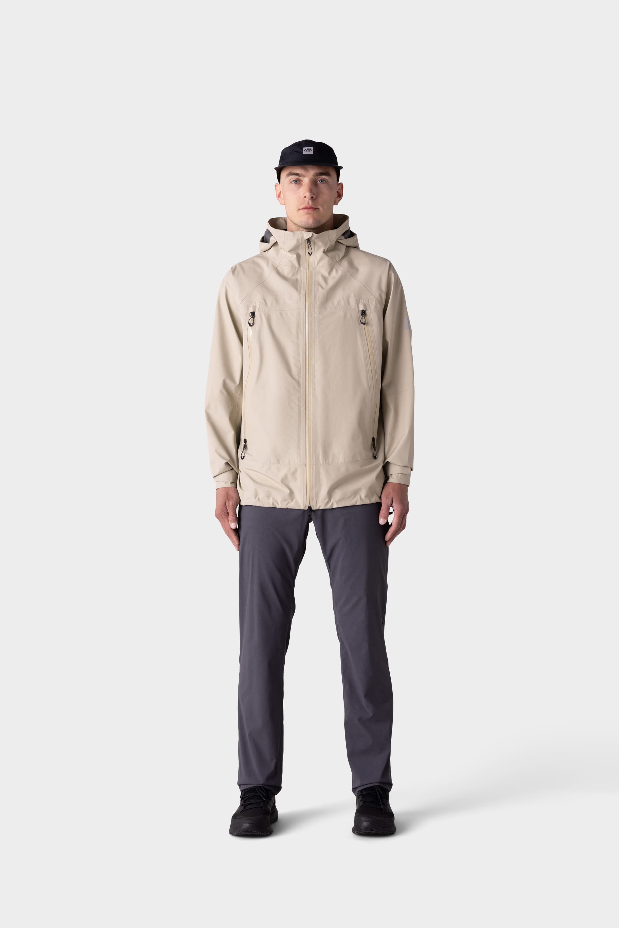 A man faces forward against a plain light background, wearing a beige hooded jacket, black baseball cap and shoes, and 686 Mens Everywhere Featherlight Chino Pant - Relaxed Fit by 686 in dark gray.