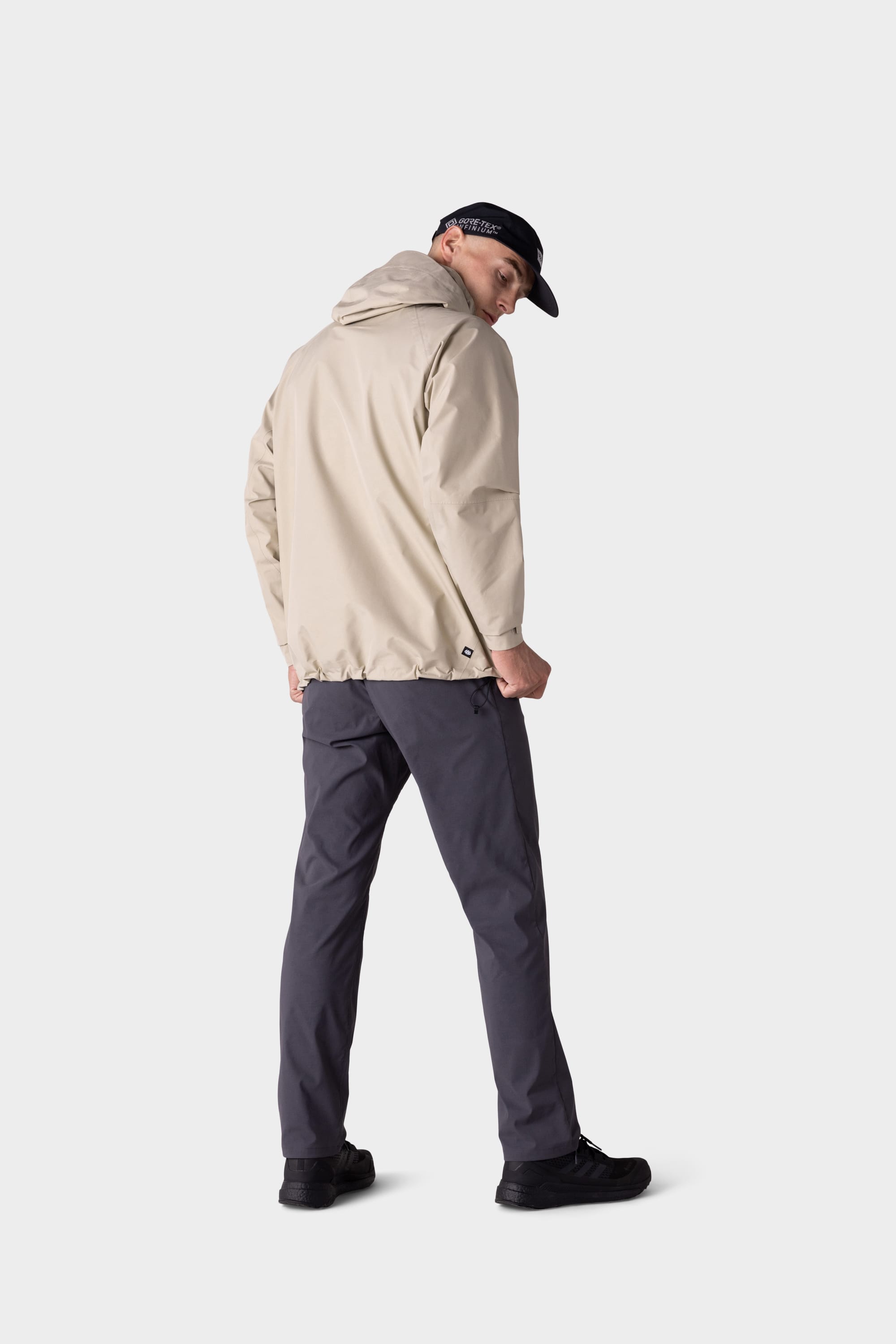 A man stands facing away, turning his head to the side against a plain light background, wearing 686 Mens Everywhere Featherlight Chino Pant - Relaxed Fit in gray, with a beige hooded jacket, black shoes, and a black cap.