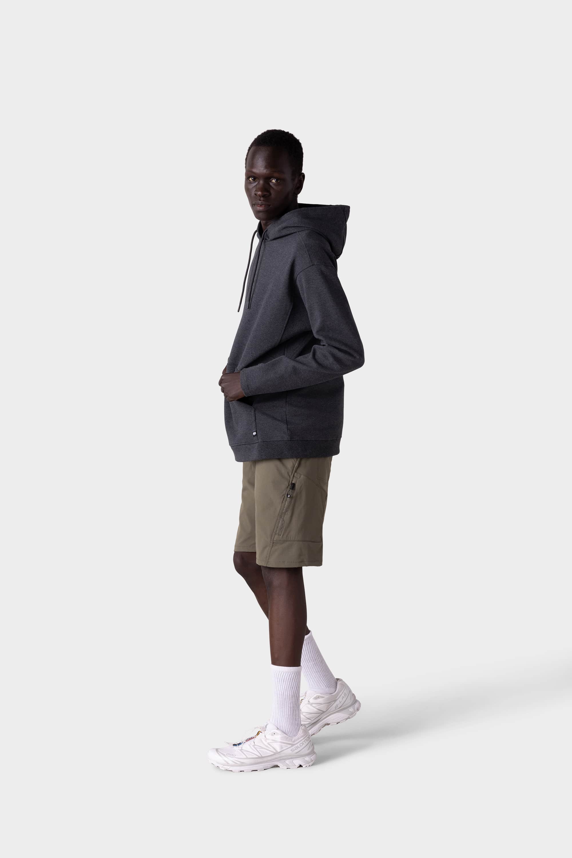 A person stands sideways against a plain background, wearing a dark gray hoodie, 686 Mens Anything Hybrid Cargo Shorts by 686 in olive green, paired with white socks and sneakers—hands in pockets, gazing at the camera in minimalist style.