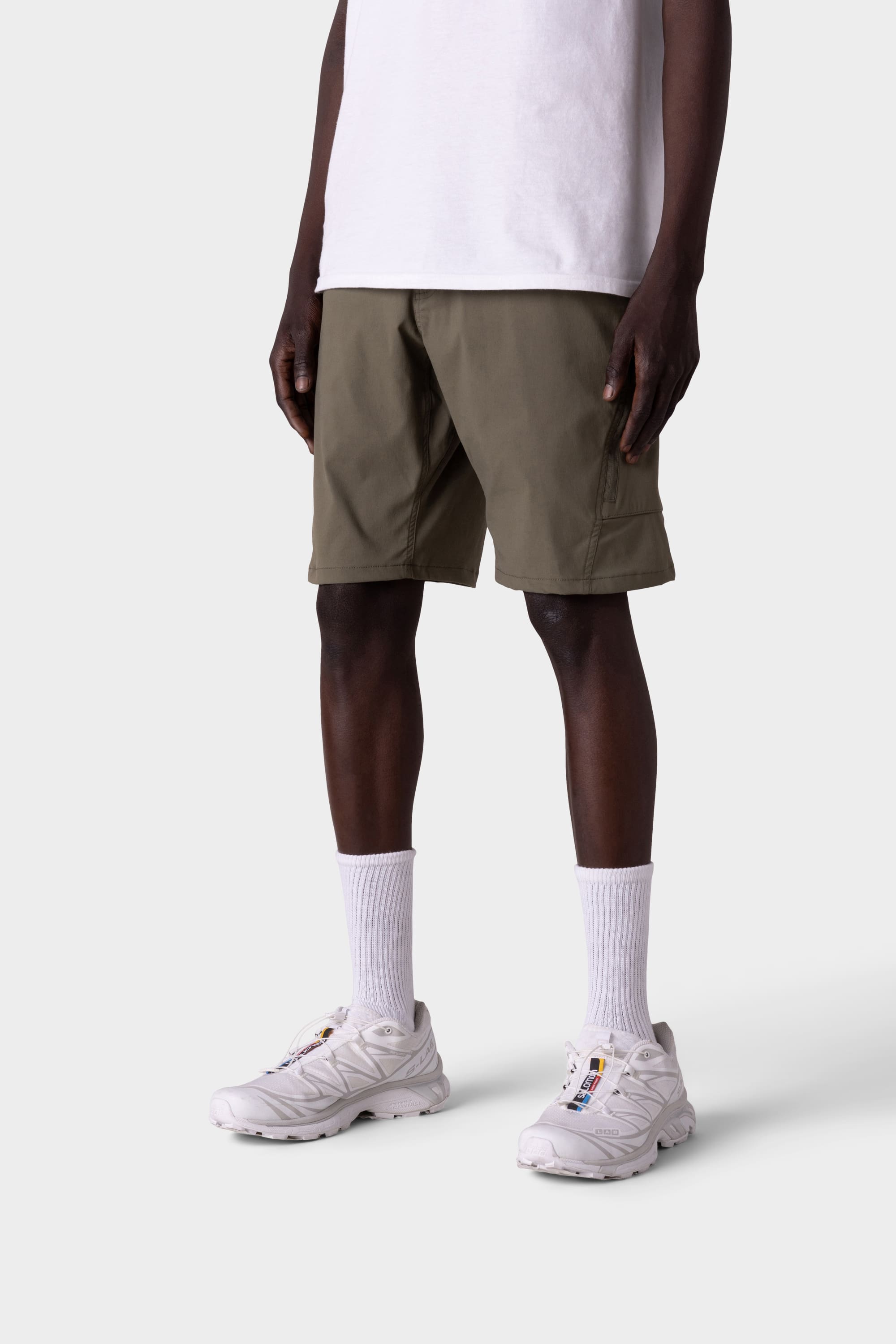 A person wears 686 Mens Anything Hybrid Cargo Short in olive green, paired with a white t-shirt, white crew socks, and white sneakers. Only the lower half is visible against a plain light gray background, emphasizing the minimalist style.