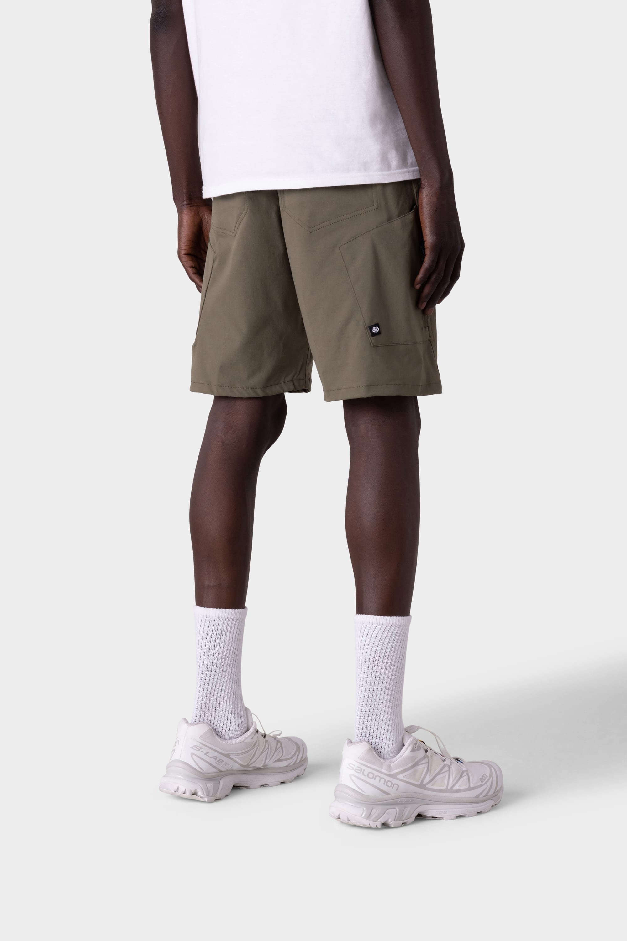 A person wears 686 Mens Anything Hybrid Cargo Short in olive green, a white T-shirt, white crew socks, and white sneakers while standing with their back to the camera against a plain light gray background.