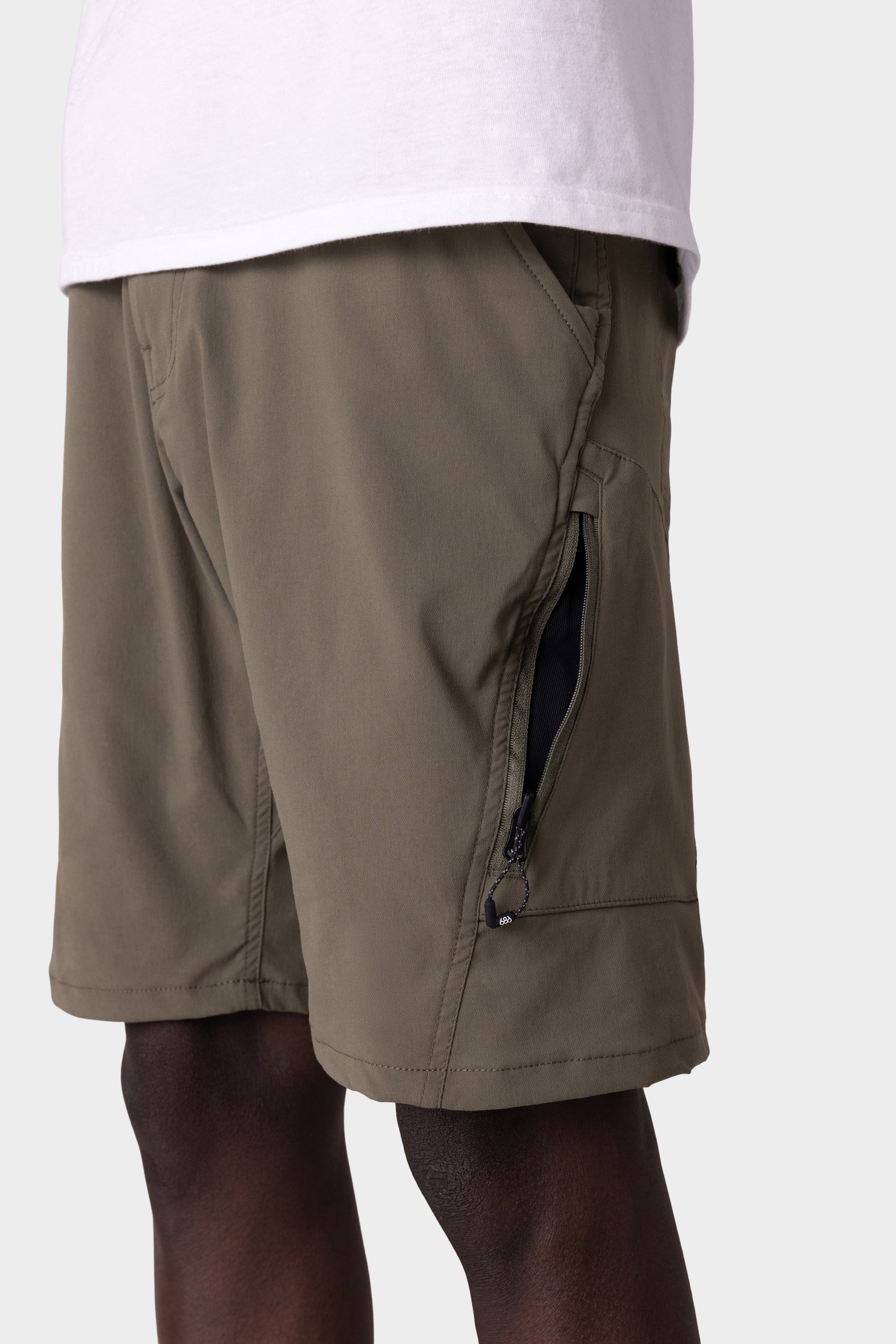 A person wears the 686 Mens Anything Hybrid Cargo Short in olive green, featuring a zippered side pocket and minimalist design, paired with a white shirt. Brand: 686.