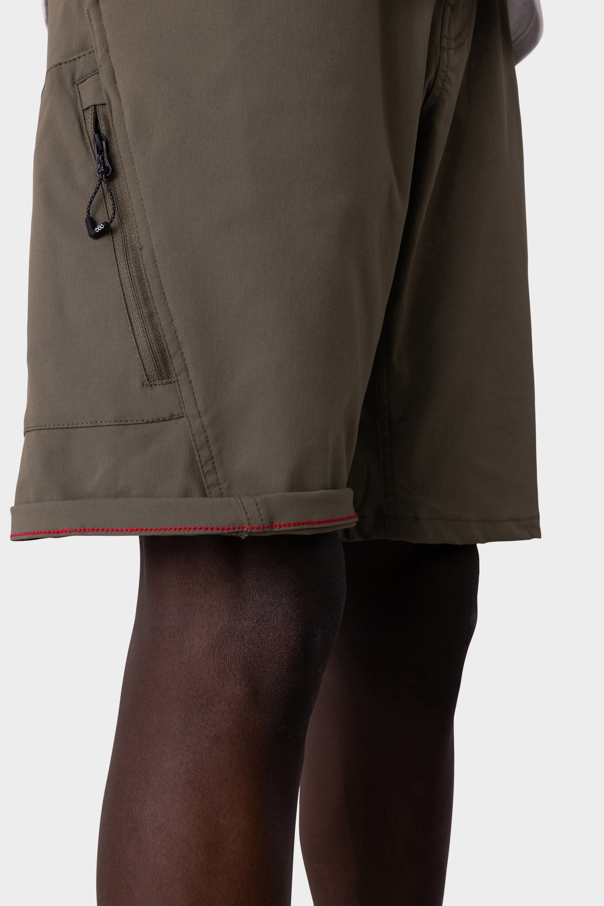 A close-up of a person wearing the 686 Mens Anything Hybrid Cargo Short by 686, featuring olive green fabric, a zippered side pocket, and red stitching at the hem, highlights the minimalist design against a light background.