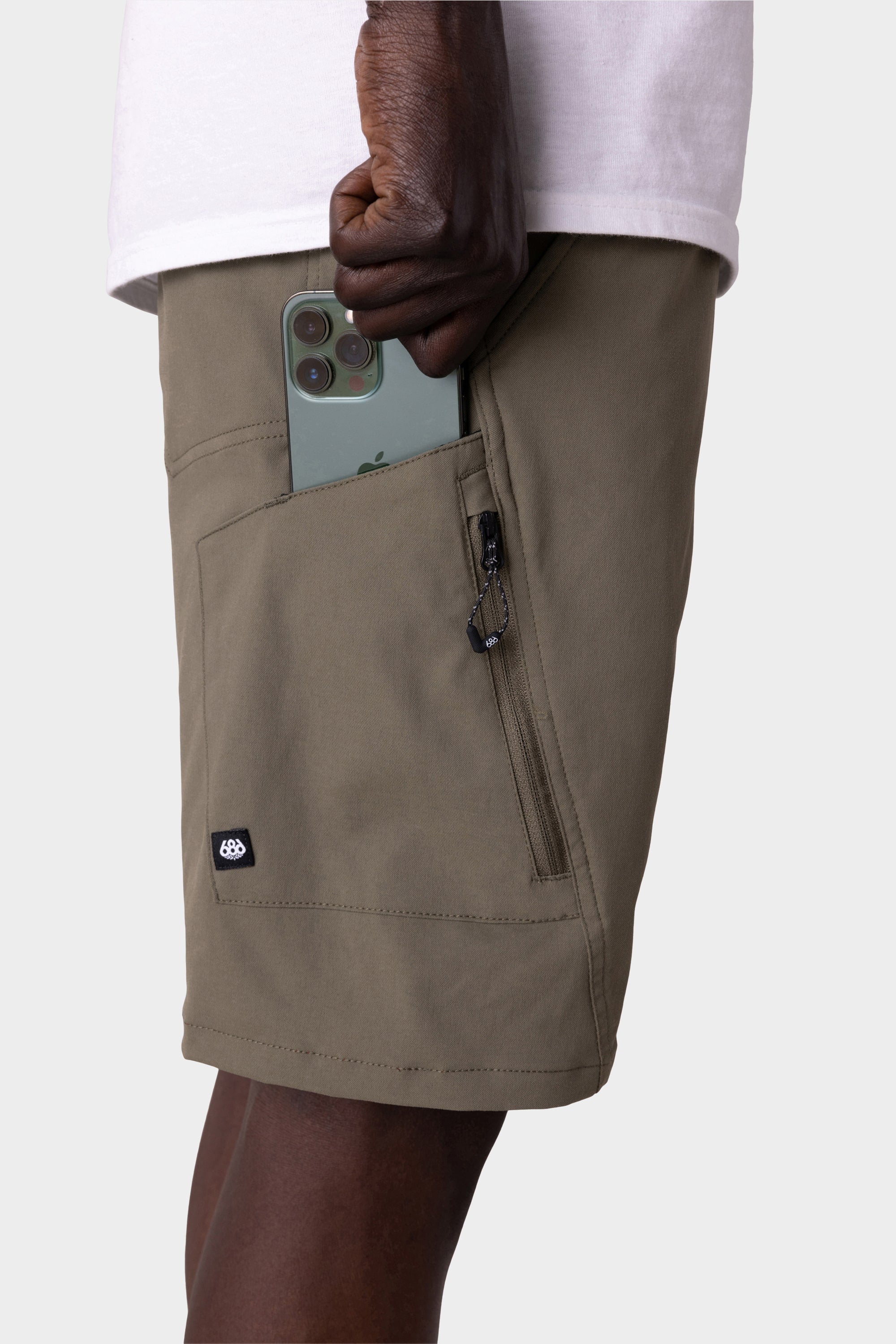 A person in 686 Mens Anything Hybrid Cargo Short by 686, featuring an olive green color and zipper pocket, places a green smartphone into the pocket while wearing a simple white shirt.