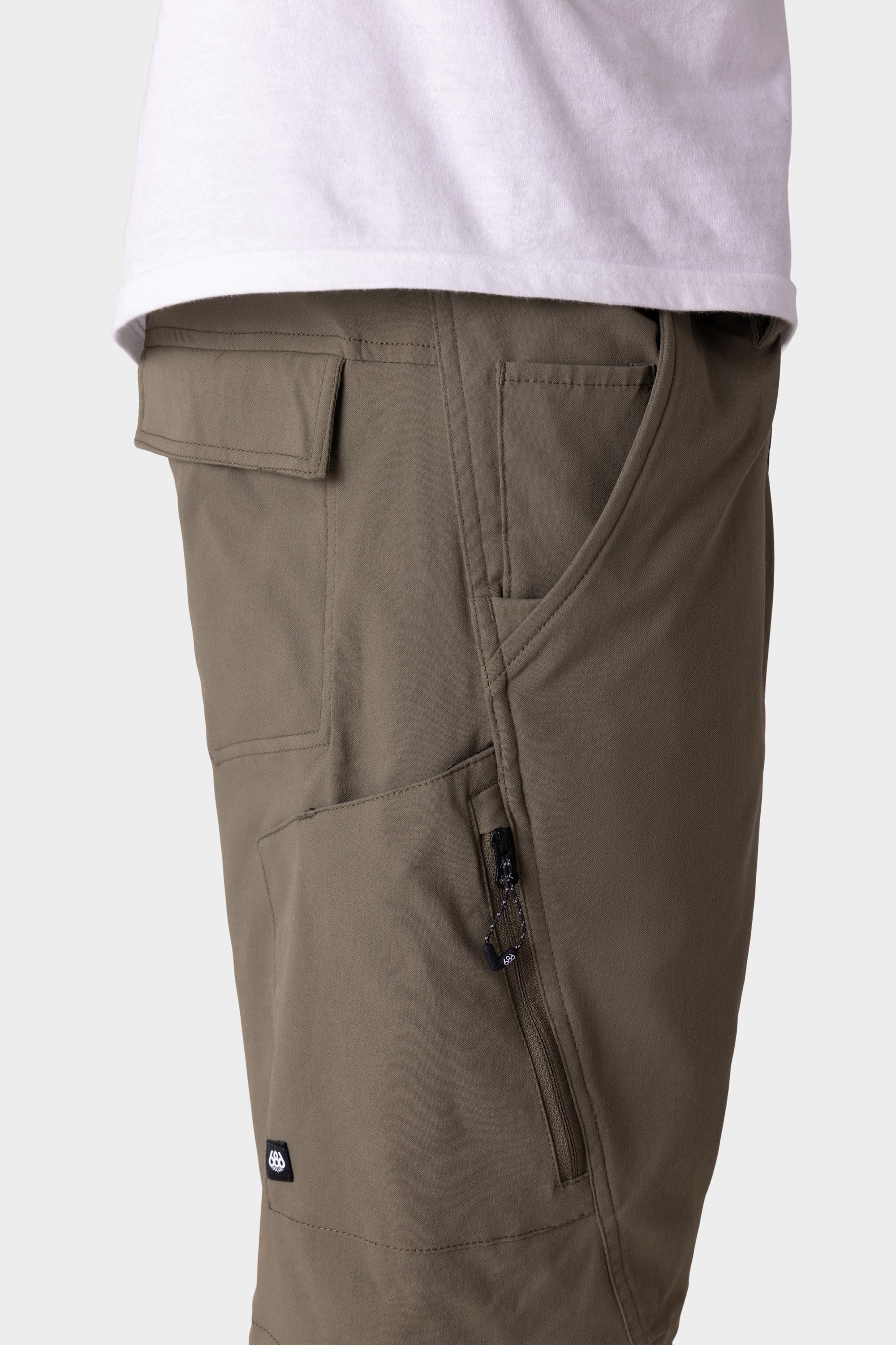 A close-up side view shows a person wearing 686 Mens Anything Hybrid Cargo Shorts by 686 in olive green, featuring multiple pockets like zippered and flap styles, paired with a white shirt for a minimalist look.