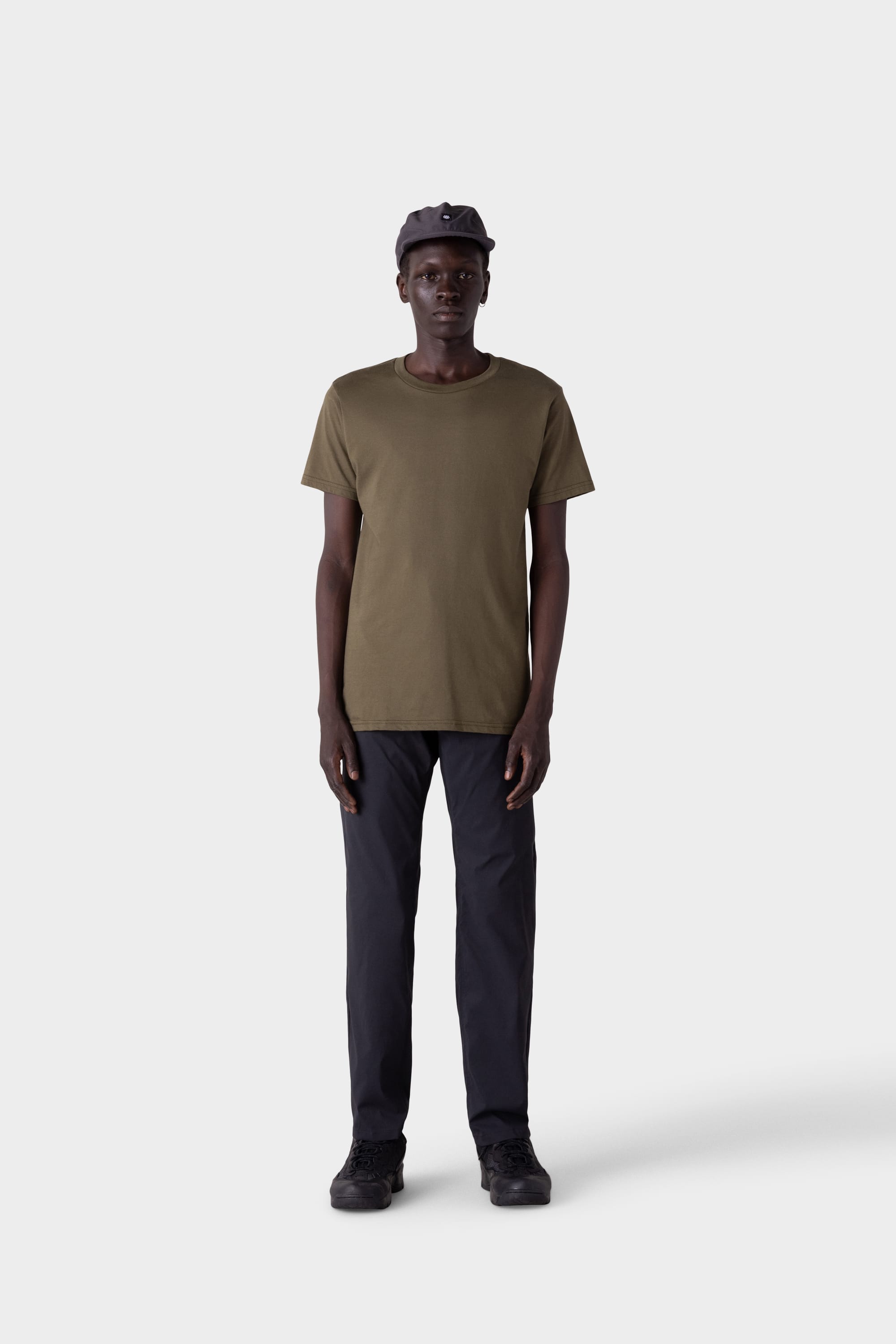 A person stands facing forward against a plain light gray background, wearing an olive green t-shirt, black shoes, a black baseball cap, and 686 Mens Everywhere Featherlight Chino Pant - Relaxed Fit by 686.