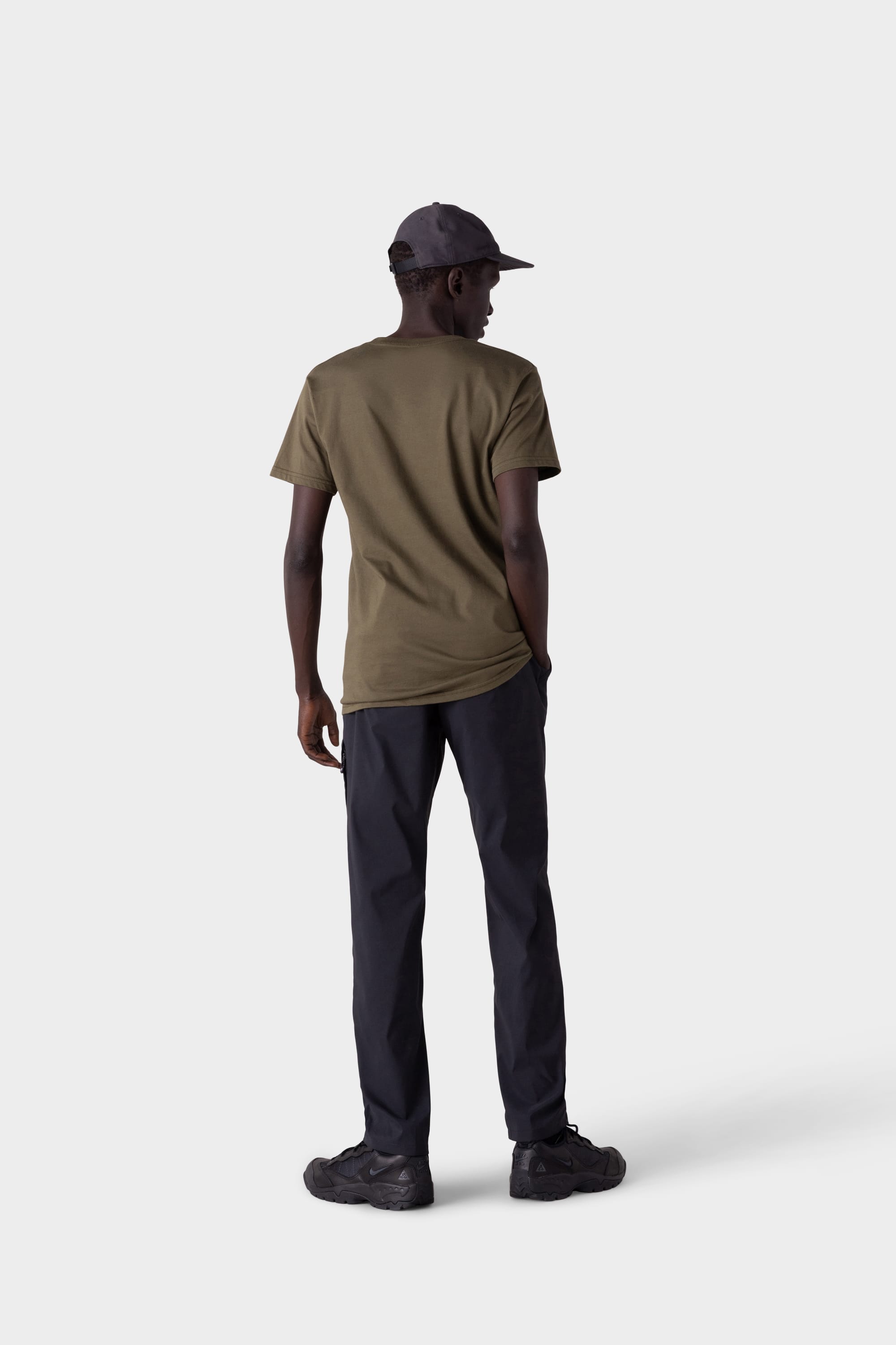 A man stands facing away against a plain light gray background, wearing an olive green t-shirt, black cap, black sneakers, and 686 Mens Everywhere Featherlight Chino Pant - Relaxed Fit by 686.