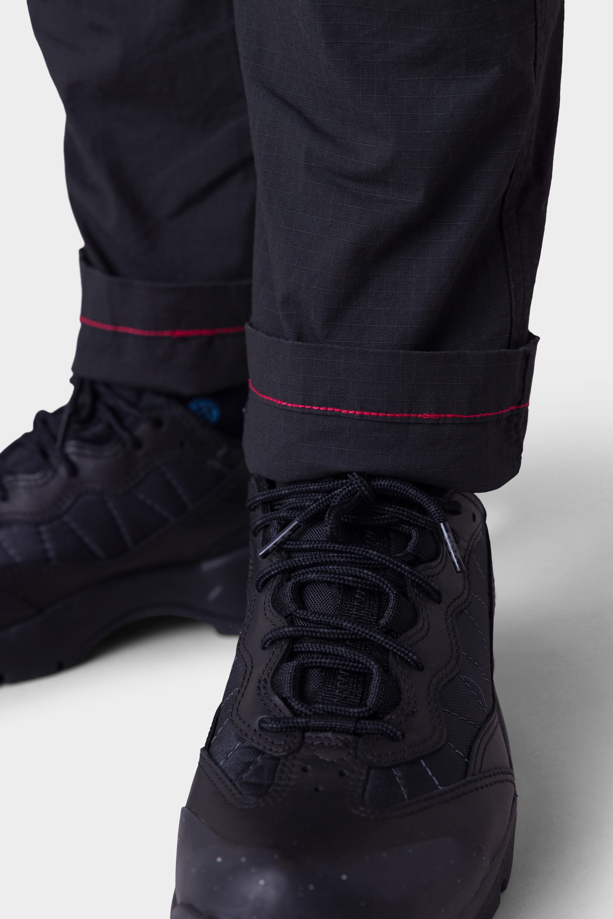 A person wears 686 Mens All Time Cargo Pant - Wide Tapered Fit with rolled-up cuffs, showing a red stitched seam, paired with black sneakers. The background is plain and light-colored.