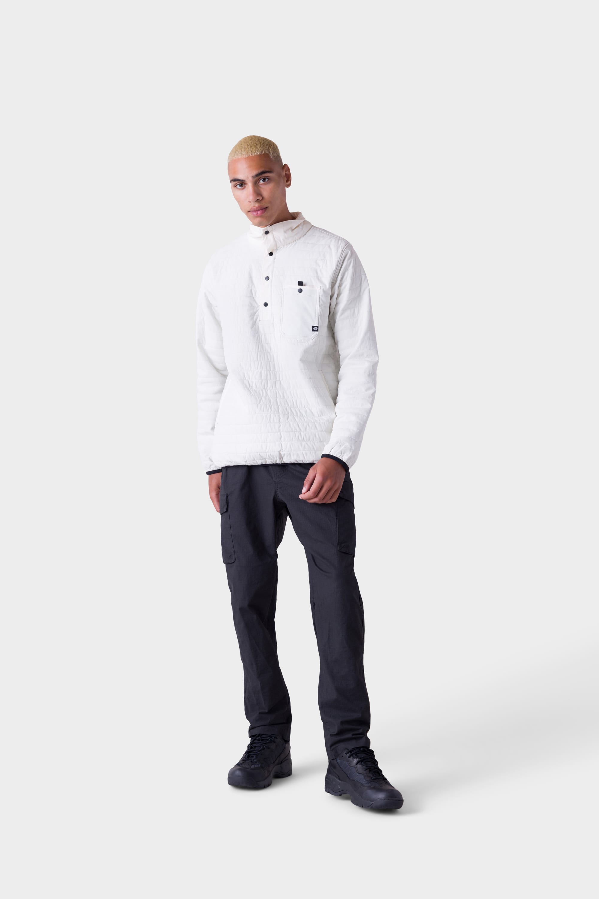A person with short blond hair in a textured white long-sleeve shirt, 686 Mens All Time Cargo Pant - Wide Tapered Fit by 686, and black shoes stands against a plain light gray background.