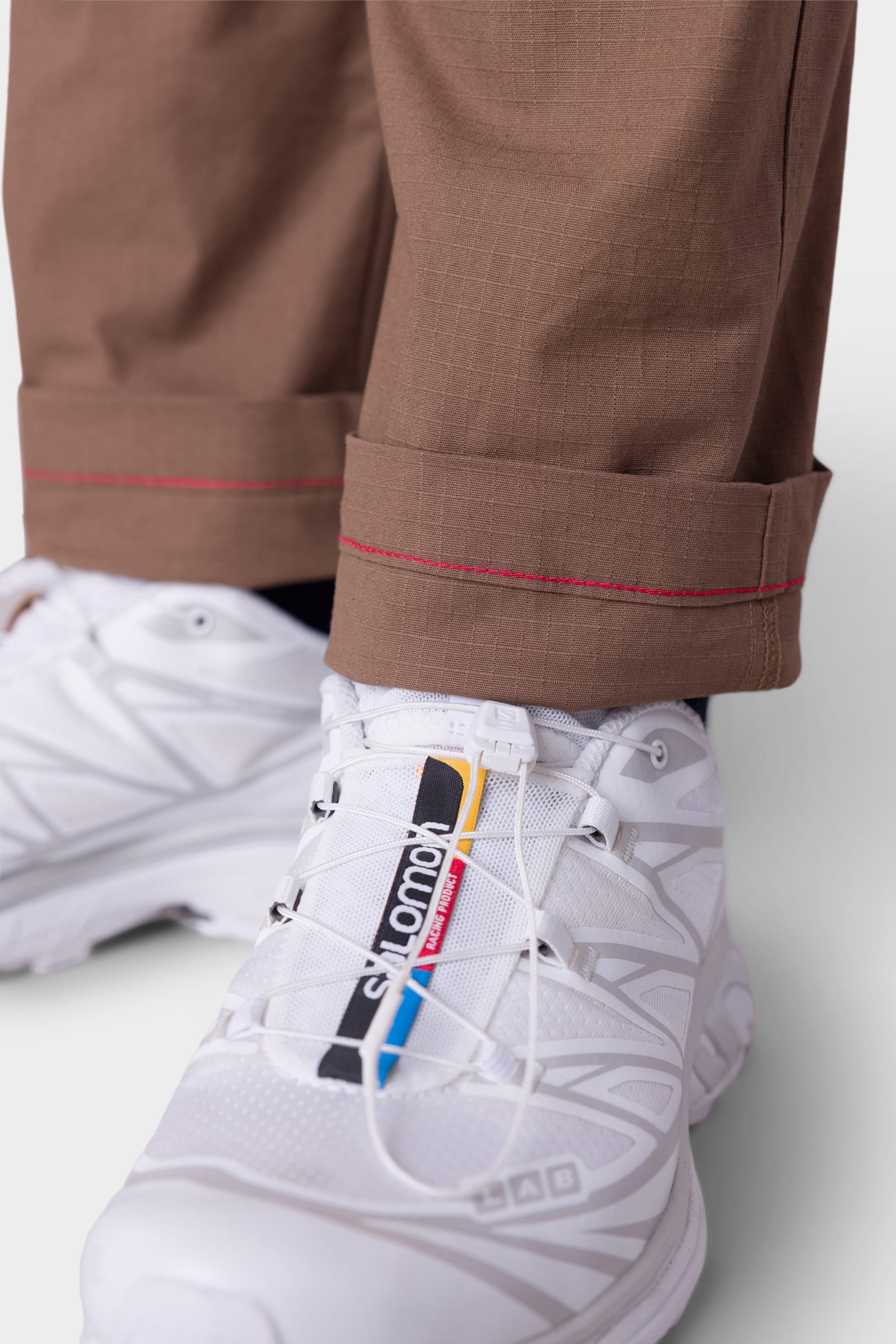 A close-up of a person wearing white Salomon sneakers with multicolored branding, paired with 686 Mens All Time Cargo Pant - Wide Tapered Fit in brown featuring red stitching near the hem.