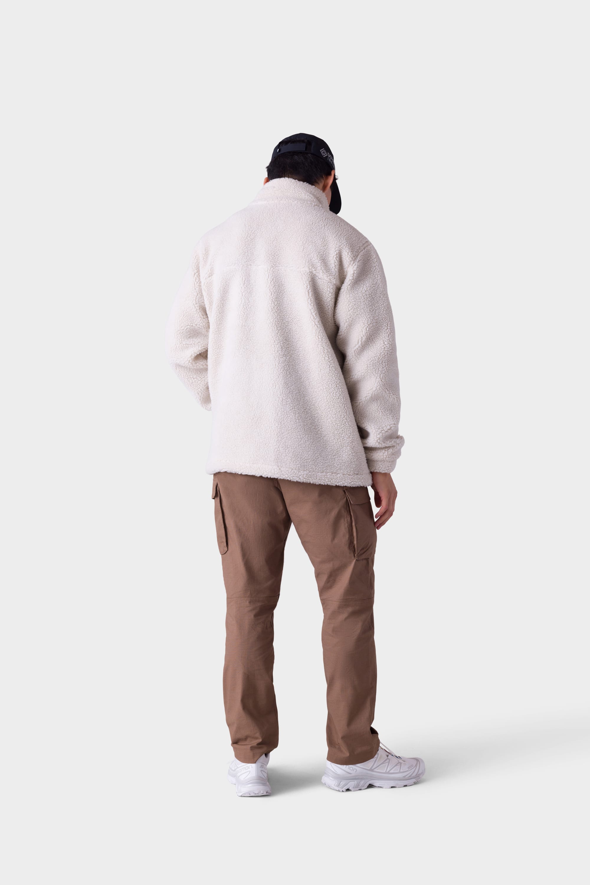 A person stands facing away against a light gray background, wearing a cream fleece jacket, white sneakers, black cap, and 686 Mens All Time Cargo Pant - Wide Tapered Fit by 686 in brown.