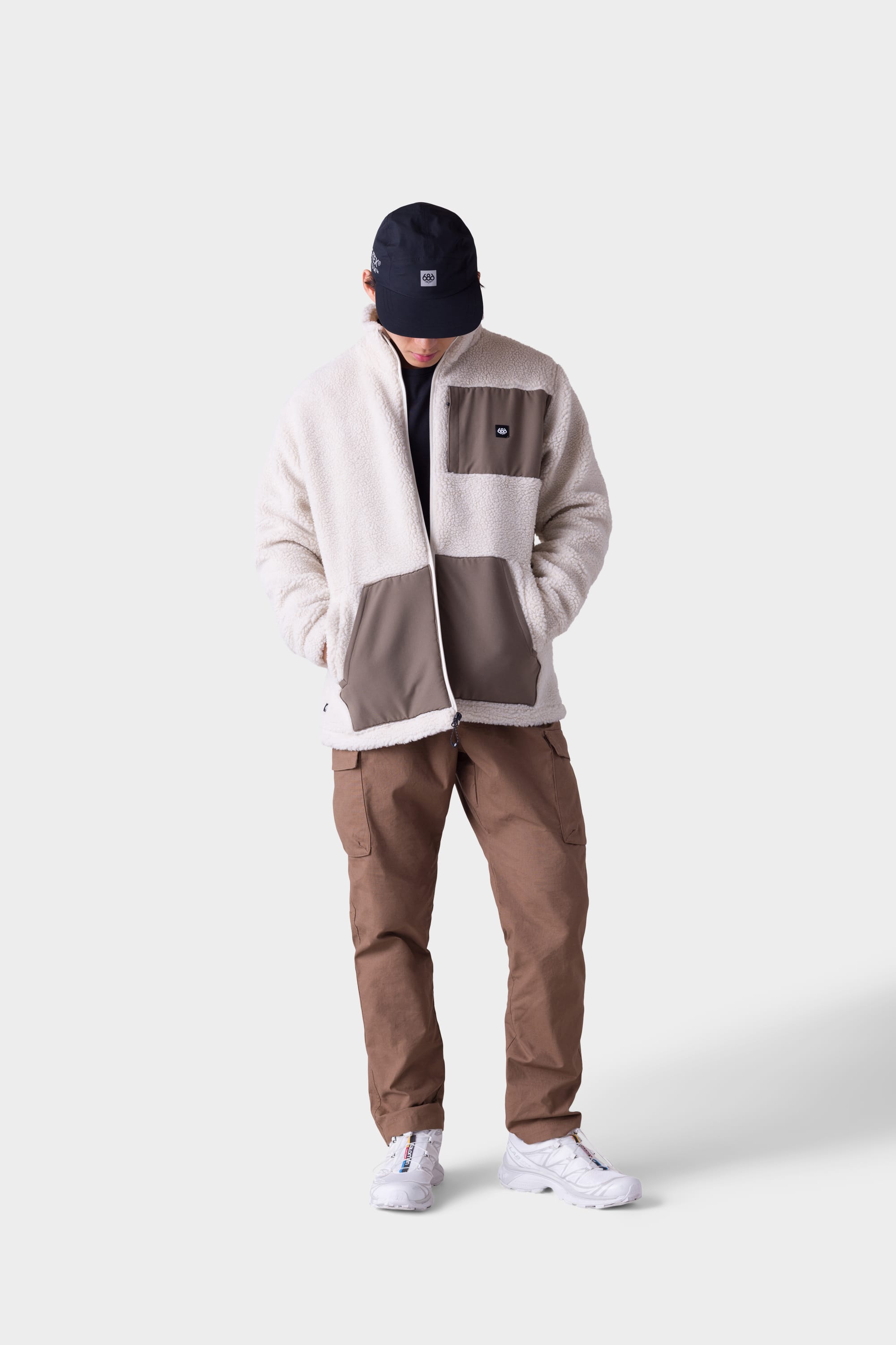 A person wearing a black cap, cream and beige sherpa jacket, 686 Mens All Time Cargo Pant - Wide Tapered Fit from 686 in brown, and white sneakers stands against a plain light background, looking downward with hands in jacket pockets.