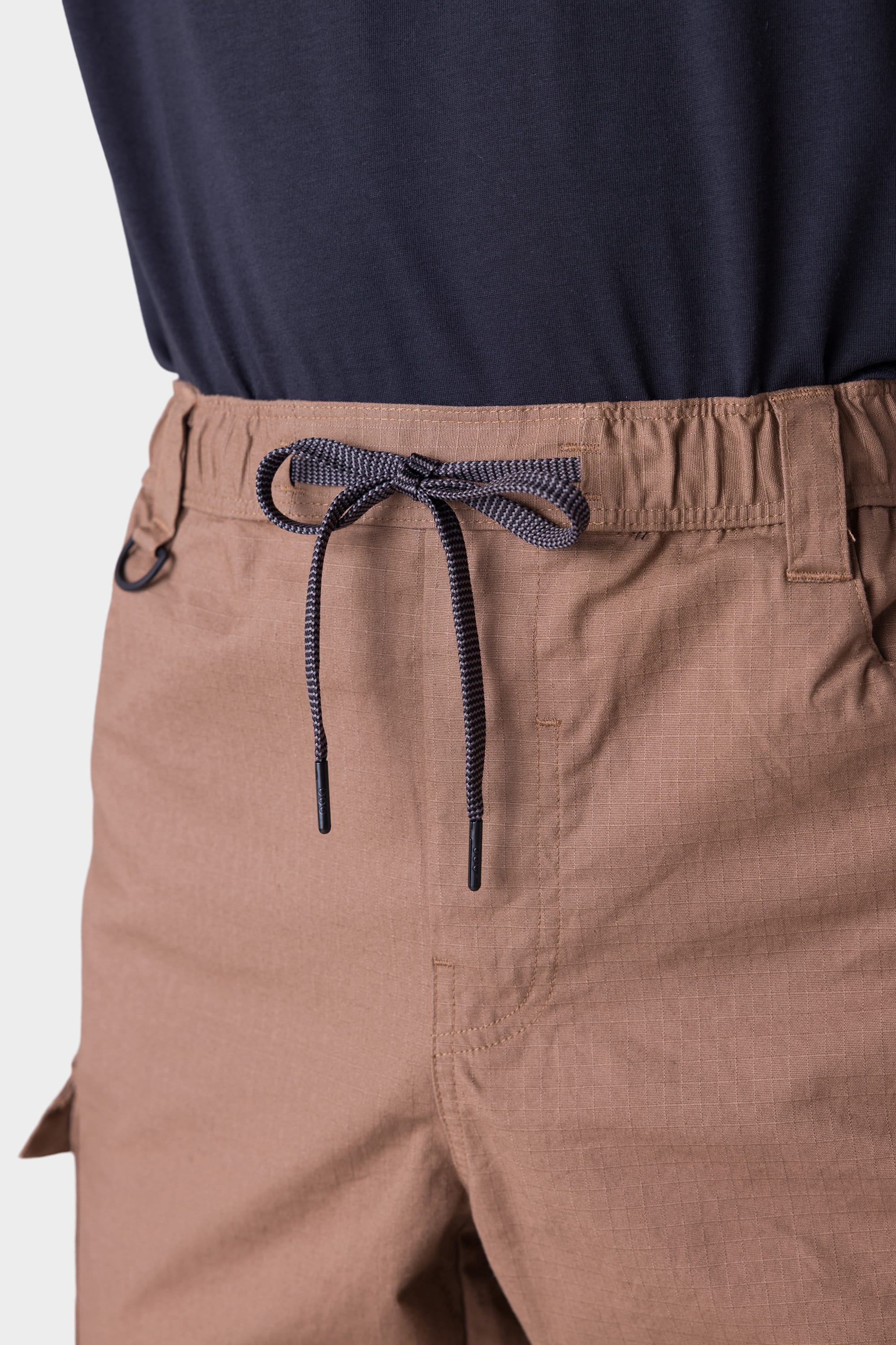 Close-up of a person wearing the 686 Mens All Time Cargo Pant - Wide Tapered Fit in light brown, featuring an elastic drawstring waist, visible stitching, and paired with a navy blue tucked-in shirt. Brand: 686.