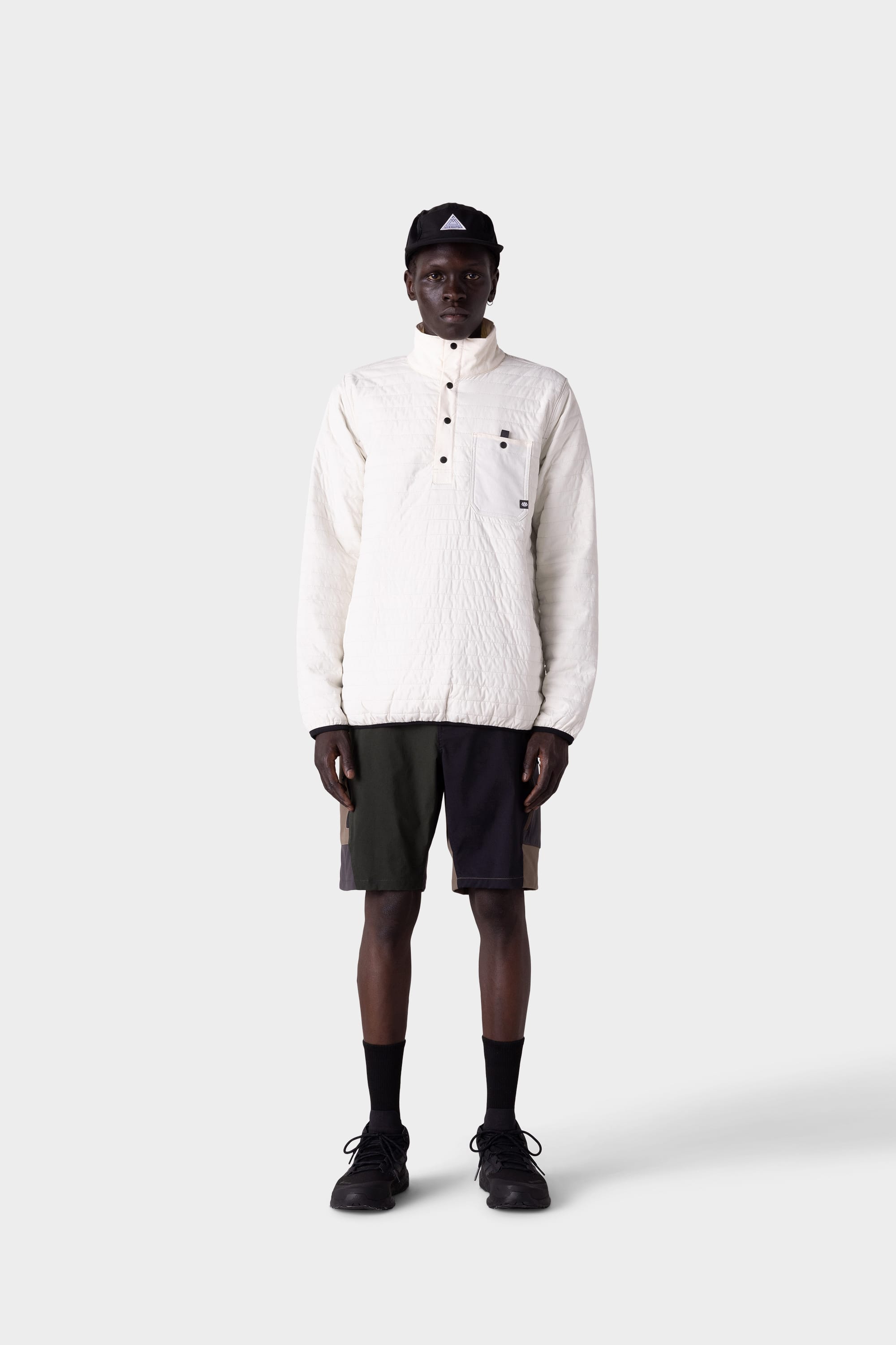 A person wearing a white quilted pullover, 686 Mens Anything Hybrid Cargo Short by 686 in black and olive green, black socks, black shoes, and a black cap stands facing forward against a plain light gray background.