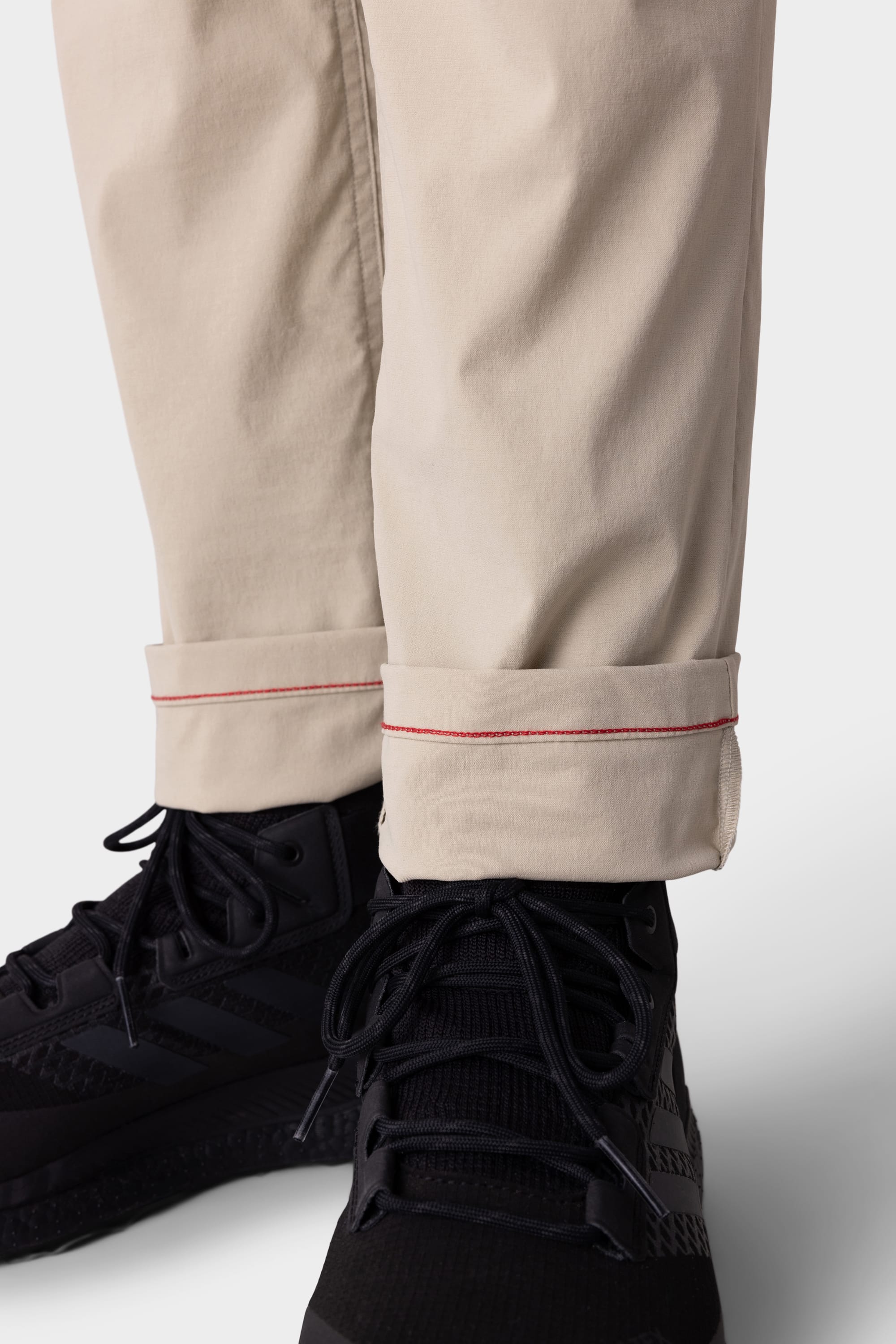 A close-up of someone wearing 686 Mens Everywhere Featherlight Chino Pant—Slim Fit, in lightweight beige with red-stitched cuffs, paired with black athletic shoes. The rolled-up style makes these chinos ideal for warm weather adventures.