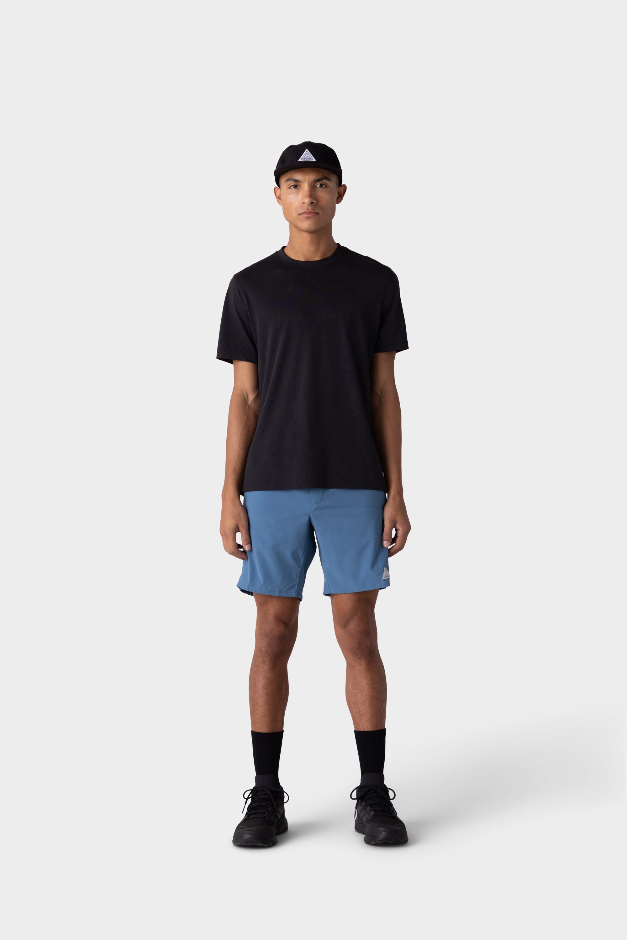 A person stands facing forward against a light gray background, wearing a black cap, black t-shirt, 686 Mens ATP Stretch Performance Short by 686 in blue, black socks, and black sneakers.